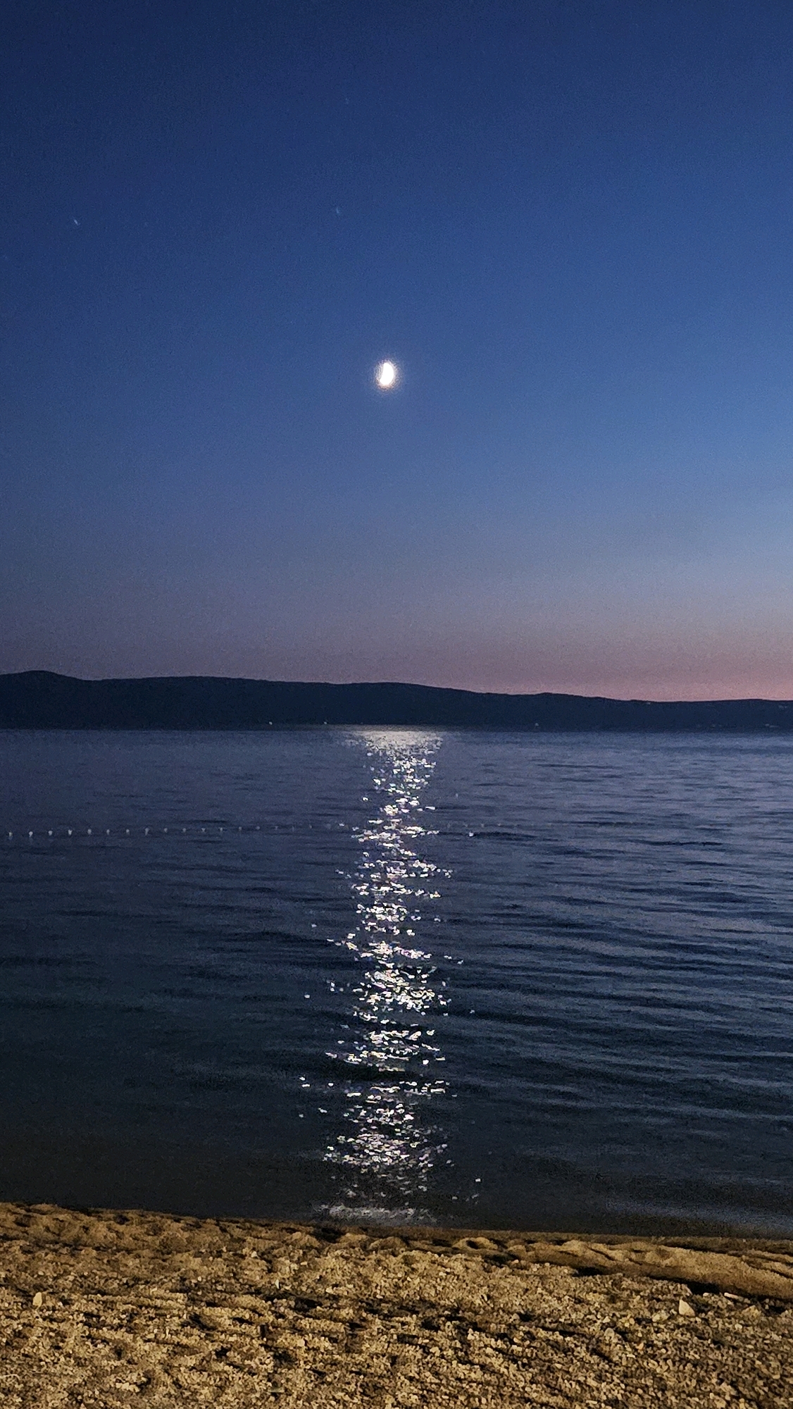 A nighttime photograph captures a serene coastal scene in Općina Tučepi, Croatia. The image is dominated by a vast expanse of deep blue sky, with a crescent moon visible near the top. Below the sky, a dark, undulating silhouette of a mountain range stretches across the horizon. In front of the mountains, the calm sea reflects the moonlight, creating a shimmering path of light across its surface. The foreground features a beach composed of pebbles and sand. The overall atmosphere is peaceful and tranquil, suggesting a quiet evening by the Adriatic Sea.