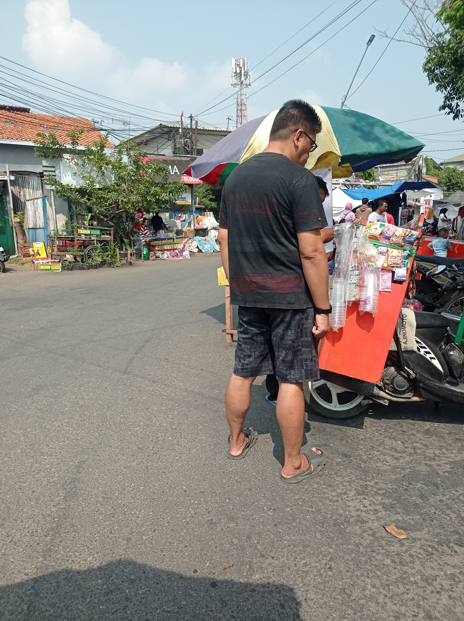 A man wearing a black t-shirt and patterned shorts stands on a paved road, his back to the camera. He is wearing flip-flops. To his right, a mobile food cart is parked, attached to the back of a motorcycle. The cart is orange and displays various packaged snacks and drinks, as well as stacks of clear plastic cups. A multi-colored umbrella, predominantly green and yellow, is open above the cart. In the background, buildings with red tile roofs are visible, along with power lines and a telecommunications tower. The scene appears to be a street-level market or roadside vendor area in Tegal, Indonesia, under a clear, bright sky.