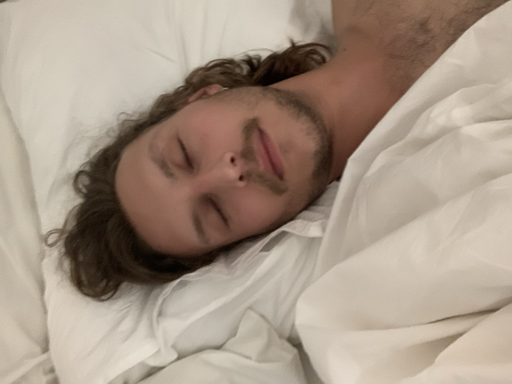 A man is asleep in bed, presumably indoors, based on the white sheets and pillows. His hair is brown and curly. He has facial hair. The location cannot be determined from the image.