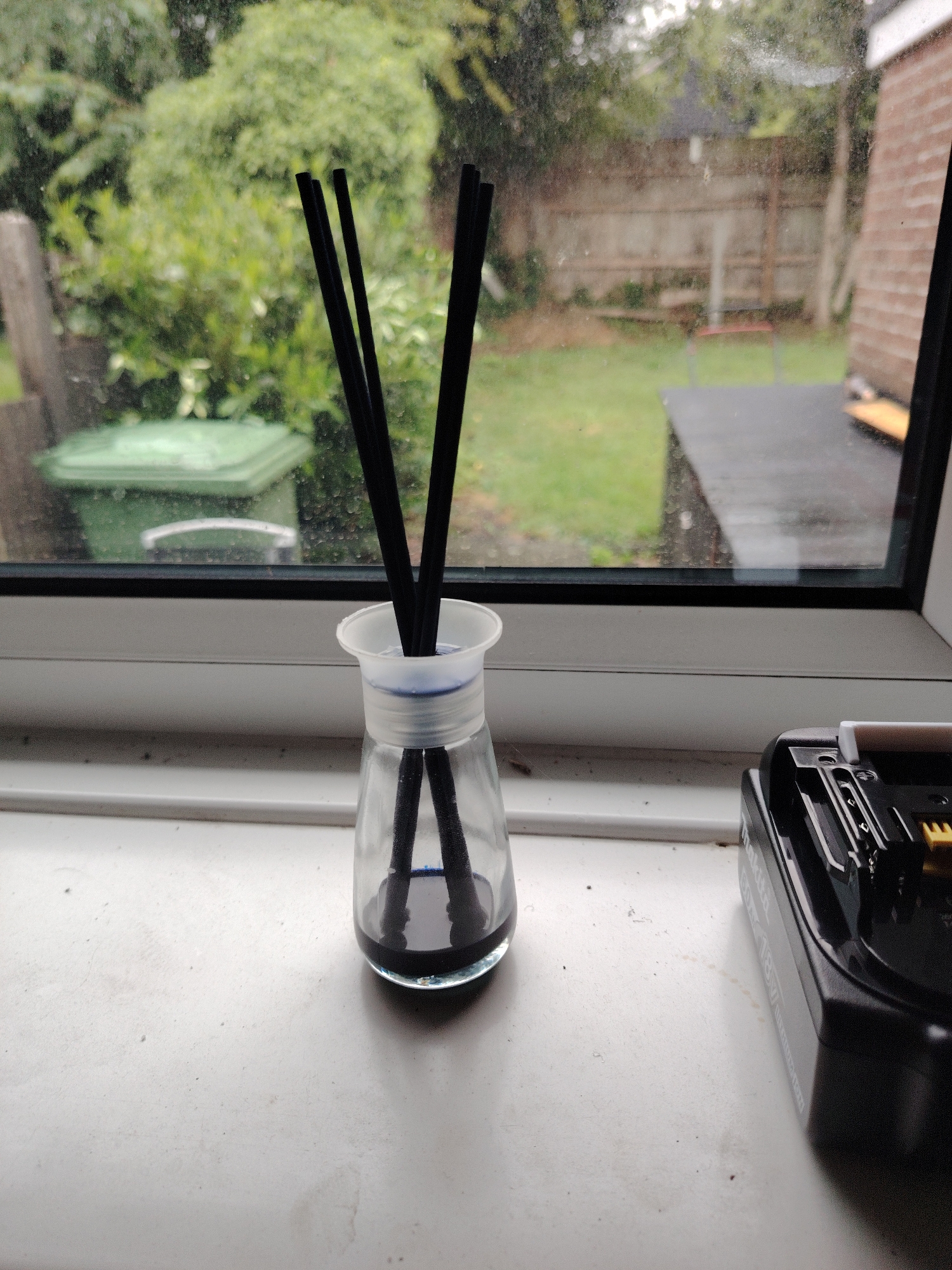 The image presents an indoor scene looking out through a window into a residential garden in Huntingdonshire, United Kingdom. In the foreground, a white window sill supports a clear glass diffuser bottle on the left, containing five black diffuser reeds submerged in a dark blue liquid, topped with a white plastic collar. To the right, a partially visible black power tool battery pack, branded 'Makita' and indicating '18V', rests on the sill. The white window frame is visible, bordered by a black seal around the glass. The window pane itself shows numerous smudges, water marks, and fine debris.
Through the window, an overcast outdoor garden is visible. Extensive green grass covers the ground, and various dense green foliage and trees are present throughout the background. A wooden fence extends horizontally across the mid-ground, delineating the property boundary. To the left, a large green wheelie bin with its lid closed is situated on the grass. Towards the right, a dark, flat outdoor surface, possibly a patio or decking, is partially visible, with a small yellow object resting upon it. A faint red object is also discernible in the distant mid-ground. No individuals are depicted within the frame.