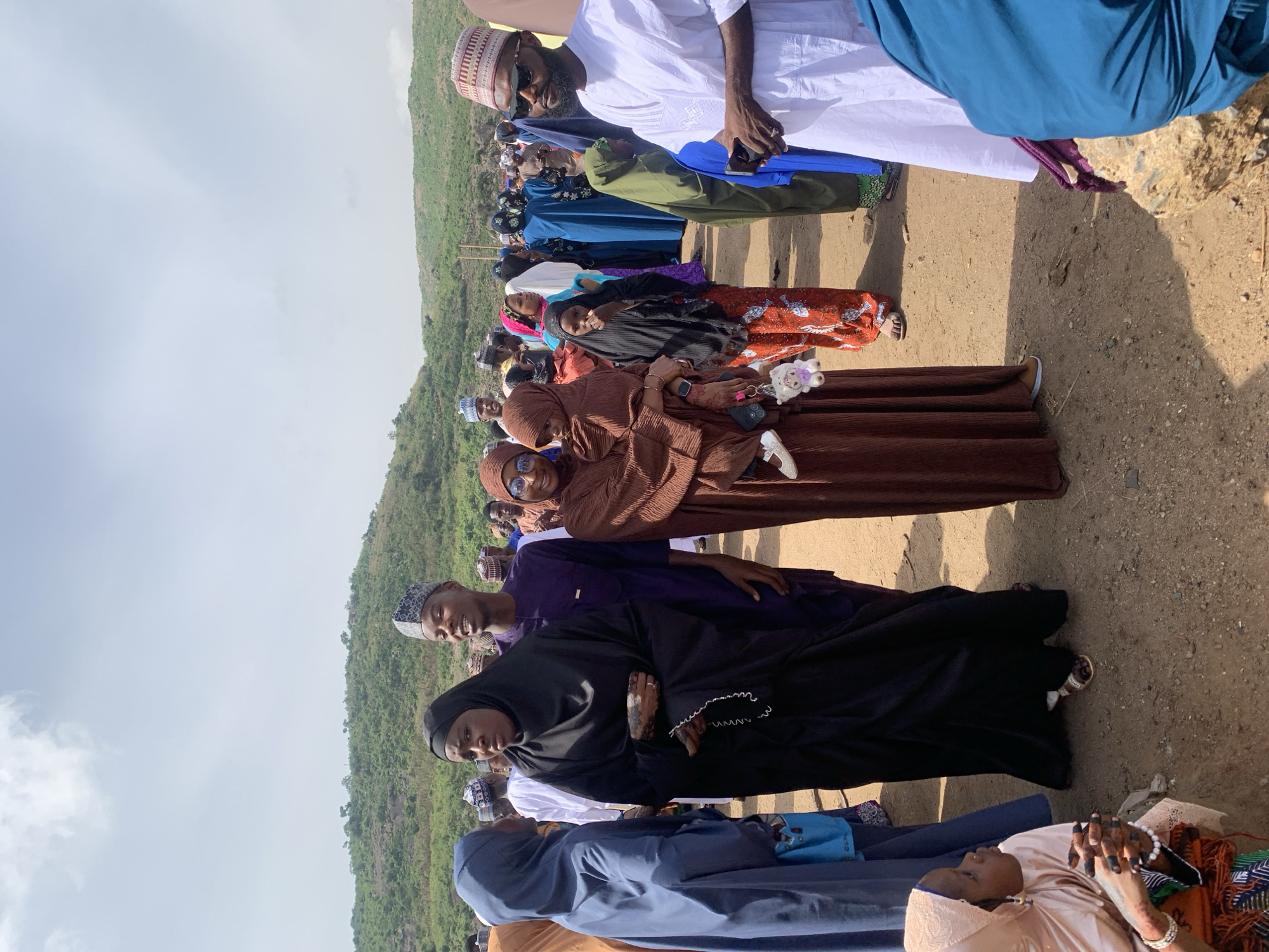 A medium-shot photograph taken outdoors shows a group of people standing or sitting on dirt ground in Naraguta Hausawa, Nigeria, during daylight hours. The scene is oriented vertically. A green, grassy hillside is in the background. A cloudy sky fills the upper portion of the frame.
Several individuals are visible, predominantly women wearing hijabs and other traditional Islamic attire. Colors range from brown and black to blue, orange, and white. A man in a white garment and cap stands at the edge of the group.
Some people are interacting while others are looking at the camera. No specific activities are readily apparent beyond the group gathering. A purse, a phone and keys are held.