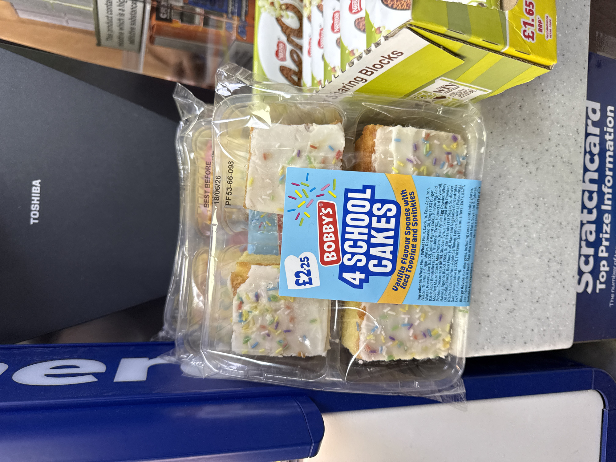 The image shows a package of "Bobby's 4 School Cakes" resting on a surface in what appears to be a retail setting in North Hertfordshire, United Kingdom. The cakes are vanilla flavored sponge cakes with iced toppings and sprinkles, packaged in a clear plastic container. A blue and white label on the package displays the product name, brand, price (£2.25), and ingredients. The cakes are likely for retail sale. The 'best before' date shown on the package is 18/06/26. Adjacent to the cakes are a box of "Sharing Blocks" and a scratchcard for Top Prize Information, also available in the area, suggesting the cakes may have been purchased at a retail location like a convenience store. A black "Toshiba" object and a blue "er" symbol are partially visible in the scene.
