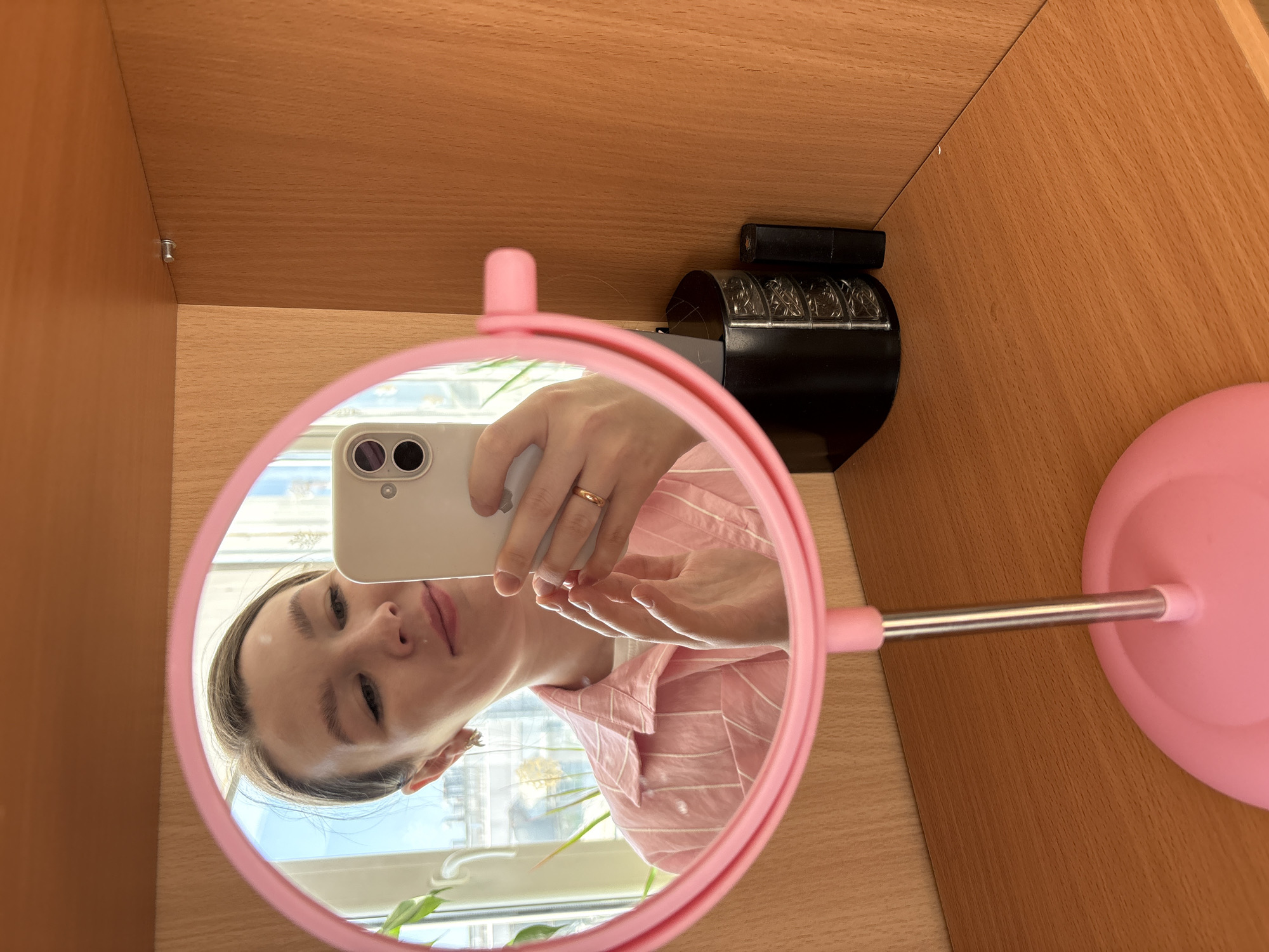 A person is taking a selfie in a pink, round mirror placed within a wooden compartment, likely a shelf or cabinet. The reflection shows the person holding a smartphone with a white case, wearing a light pink and white striped shirt, and a ring on their finger. The background reflected in the mirror shows a window, greenery, and what appears to be an urban landscape. The mirror is positioned on a wooden surface. To the side of the mirror is a black object with a series of numerical or patterned wheels. The wood grain suggests a manufactured veneer finish commonly found in furniture. There is no definitive information to confirm the location is Izhevsk, Russia from the provided image.