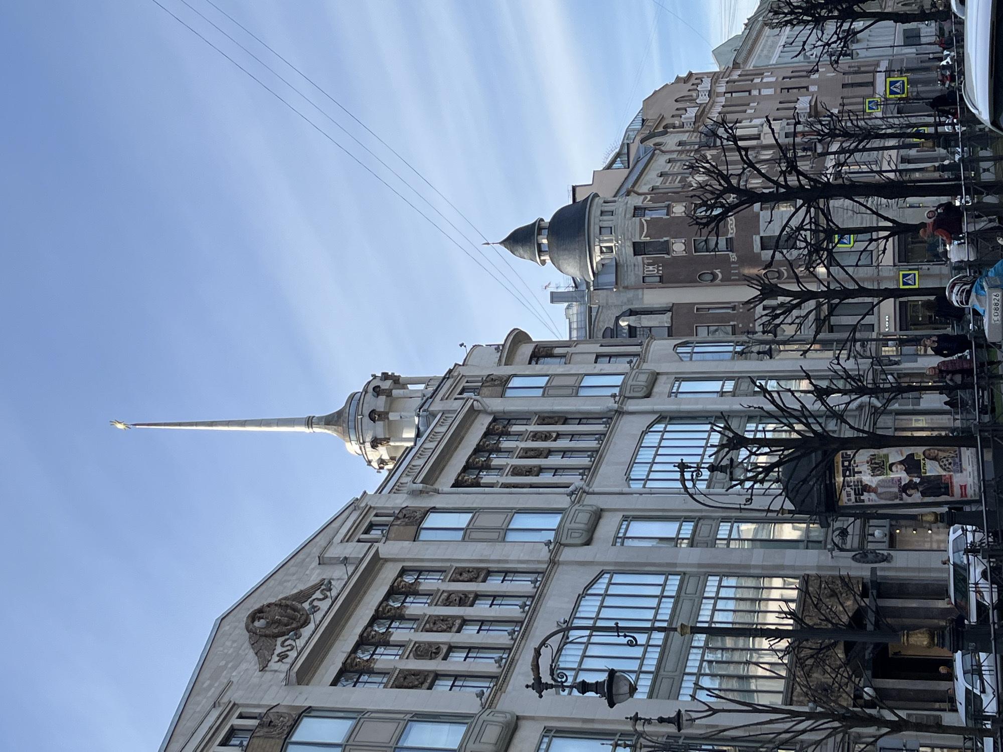 The image depicts the Singer House, located on Nevsky Prospekt in Saint Petersburg, Russia. The building is a prominent architectural landmark. The image captures the ornate facade of the Singer House, with its numerous windows and sculptural details. A spire is visible atop the building. The photograph is taken from street level, showing trees with bare branches lining the sidewalk. Pedestrians and parked vehicles are also visible on the street. The sky is blue with wispy clouds.