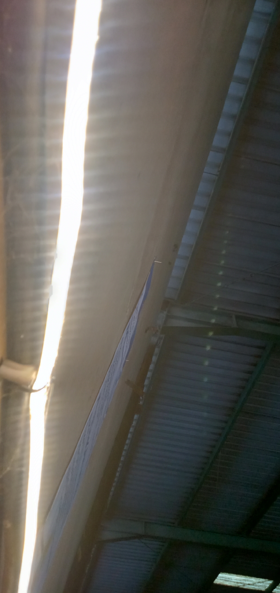 The image presents a perspective of a roof interior, likely of an industrial or commercial structure, possibly located in Banjaran, Indonesia, although location information isn't directly evident in the image. The roof construction features corrugated metal sheets supported by a framework of green-painted steel beams. A prominent, bright strip of light penetrates through a gap in the roof, casting strong shadows and highlighting the corrugated pattern. A narrow, possibly insulated panel runs along one side of the light gap. The angle suggests the photo was taken looking upwards towards the roof. No individuals, specific objects, or actions can be directly identified within the image besides the architectural elements.