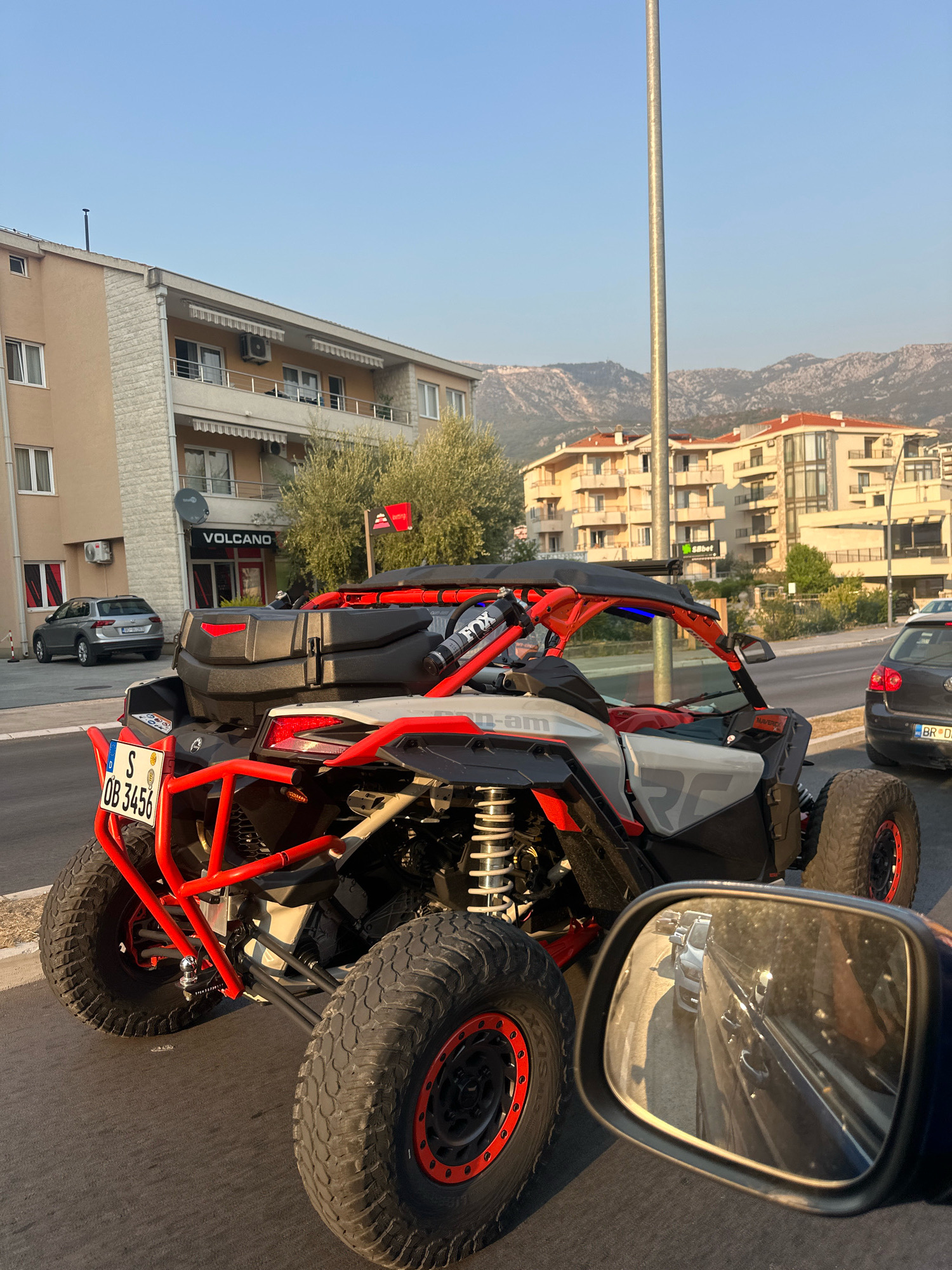 The image depicts a side view of a red and white Can-Am Maverick X3 UTV parked on a paved road in Boreti, Montenegro. The UTV has large off-road tires and a roll cage. A black storage box is mounted on the rear. To the left of the UTV, a silver Volkswagen hatchback is parked near a beige building with several windows and balconies. Above the building's entrance, a sign reads "VOLCANO." In the background, there are residential buildings with red roofs and a mountain range. A side mirror of a vehicle, likely the one from which the photo was taken, is visible in the foreground, reflecting other cars on the road. The sky is clear and blue, indicating daytime.