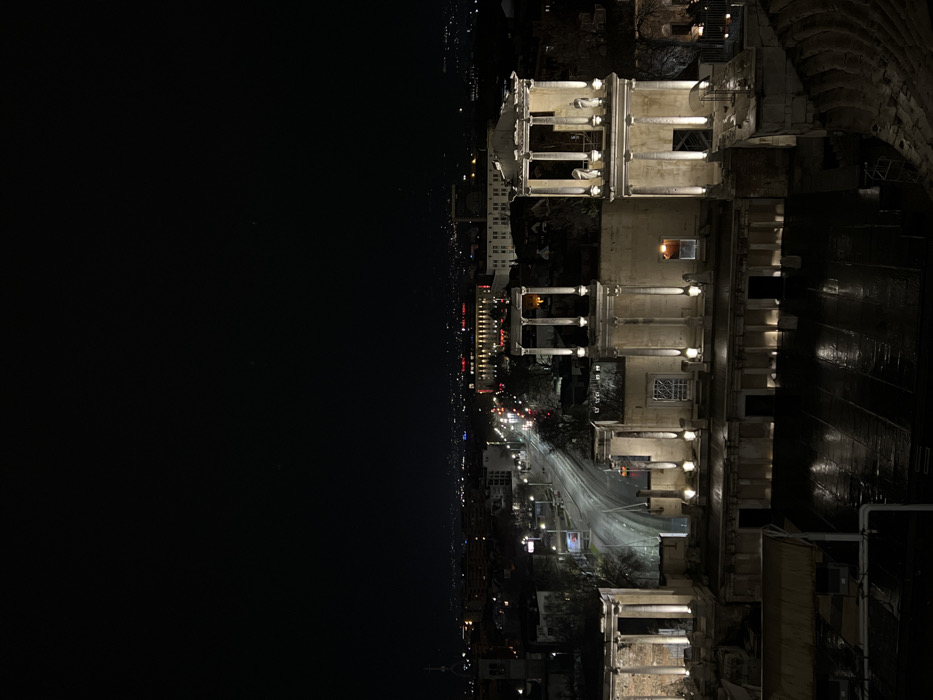 The image depicts a nighttime panoramic view of the Roman Stadium of Philippopolis in Plovdiv, Bulgaria, observed from an elevated perspective. The scene is illuminated by artificial light against a dark, clear sky.
In the mid-ground, the preserved northern facade of the Roman Stadium dominates, comprising multi-level stone structures with numerous columns and arched openings. These ancient ruins are extensively lit by warm-toned spotlights positioned at their base and along vertical architectural elements, highlighting the details of the beige stone, architraves, and the statuary visible on the upper level of the rightmost section. The ground level directly in front of these structures appears to be a dark, reflective surface, possibly wet paving.
To the left of the Roman Stadium facade, a city street, identified as Knyaz Alexander I Street, curves towards the background. The street is illuminated by streetlights, and streaks of red taillights and white headlights indicate active vehicle traffic. Buildings with illuminated windows line both sides of the street. Further into the background, the sprawling urban landscape of Plovdiv extends, marked by a multitude of distant city lights and the silhouettes of various buildings.
In the bottom right corner of the foreground, a section of dark, tiered stone seating, characteristic of an ancient stadium or amphitheater, is partially visible. No individuals are discernible within the immediate foreground or on the ancient structures. The overall environment is urban and historical, presented at night under artificial illumination.