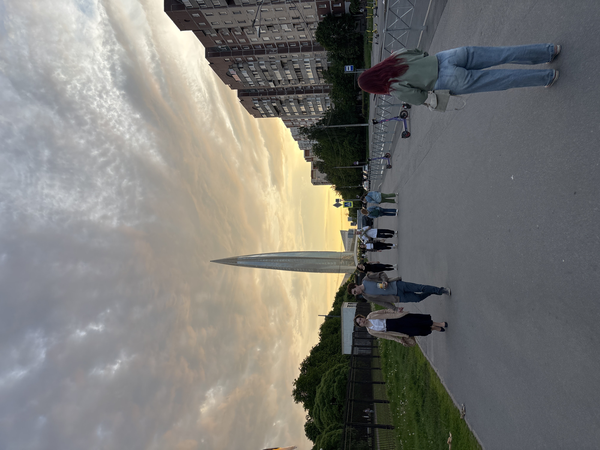 This outdoor urban scene, likely captured during sunset or sunrise, depicts a promenade in Saint Petersburg, Russia. The image is oriented vertically (rotated 90 degrees clockwise).
The sky dominates the upper portion, showcasing dramatic clouds in a mix of warm golden-orange hues on the right, transitioning to cooler grays and whites with textured patterns on the left, indicative of low sun angles.
The prominent subject is the slender, glass-clad Lakhta Center skyscraper, rising sharply in the middle ground. To its left, a row of older, multi-story residential buildings with numerous windows and balconies lines the street.
A wide paved road or sidewalk stretches through the foreground and mid-ground. It's bordered by a strip of green grass with small white flowers, enclosed by a metal fence. Several individuals are present, walking or standing. In the foreground, a person with reddish hair, wearing a green jacket and blue jeans, walks away from the viewer. Further back, a man in a brown jacket holds a cup, alongside a woman in a beige jacket and black skirt. Other figures are visible in the distance.
Along the roadside barrier, multiple electric scooters are lined up, suggesting a rental service. Street signs, including a blue bus stop sign (possibly with the Cyrillic letter 'A') and a pedestrian crossing symbol, are visible.
The scene captures a peaceful moment in a modern Russian city, blending contemporary architecture with a bustling public space under a picturesque sky. No strong emotions are discernible from the individuals.