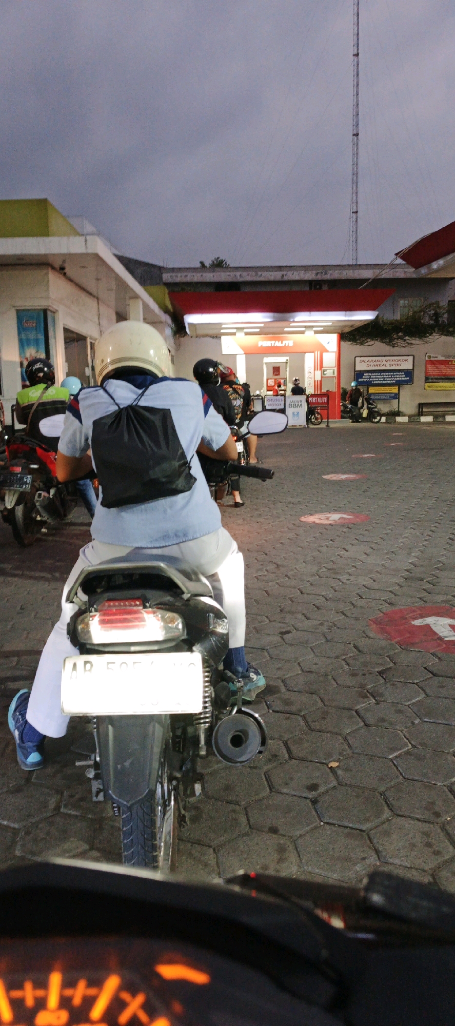 The image depicts a scene at a Pertamina gas station in Kasihan, Indonesia, during the late afternoon or early evening. The focus is on the rear view of a person sitting on a motorcycle, wearing a light blue short-sleeved shirt, white pants, a white helmet, and a black drawstring backpack. The motorcycle has a white license plate, partially obscured. Other motorcycles and people are visible in the background, some queuing at the gas pumps. The gas station canopy, with "PERTAMALITE" branding, is illuminated, and signage indicating "KHUSUS MOTOR" (for motorcycles) and "JALUR BBM" (fuel lane) is present. The ground is paved with hexagonal interlocking stones, marked with red circular indicators. A tall communications tower is visible in the distance against a cloudy sky. The overall atmosphere suggests a routine activity at a public service facility.