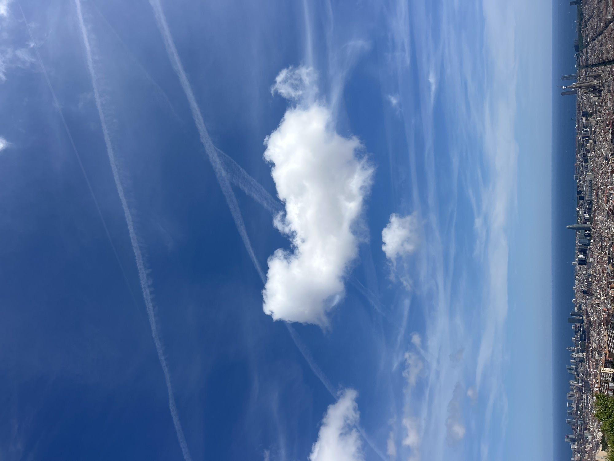 The image depicts a bright blue sky with scattered white clouds and contrails. A dense cityscape is visible on the right edge of the image, bordering the sea. The city is Barcelona, Spain, seen from an elevated vantage point, possibly Montjuïc Hill. No people are visible.