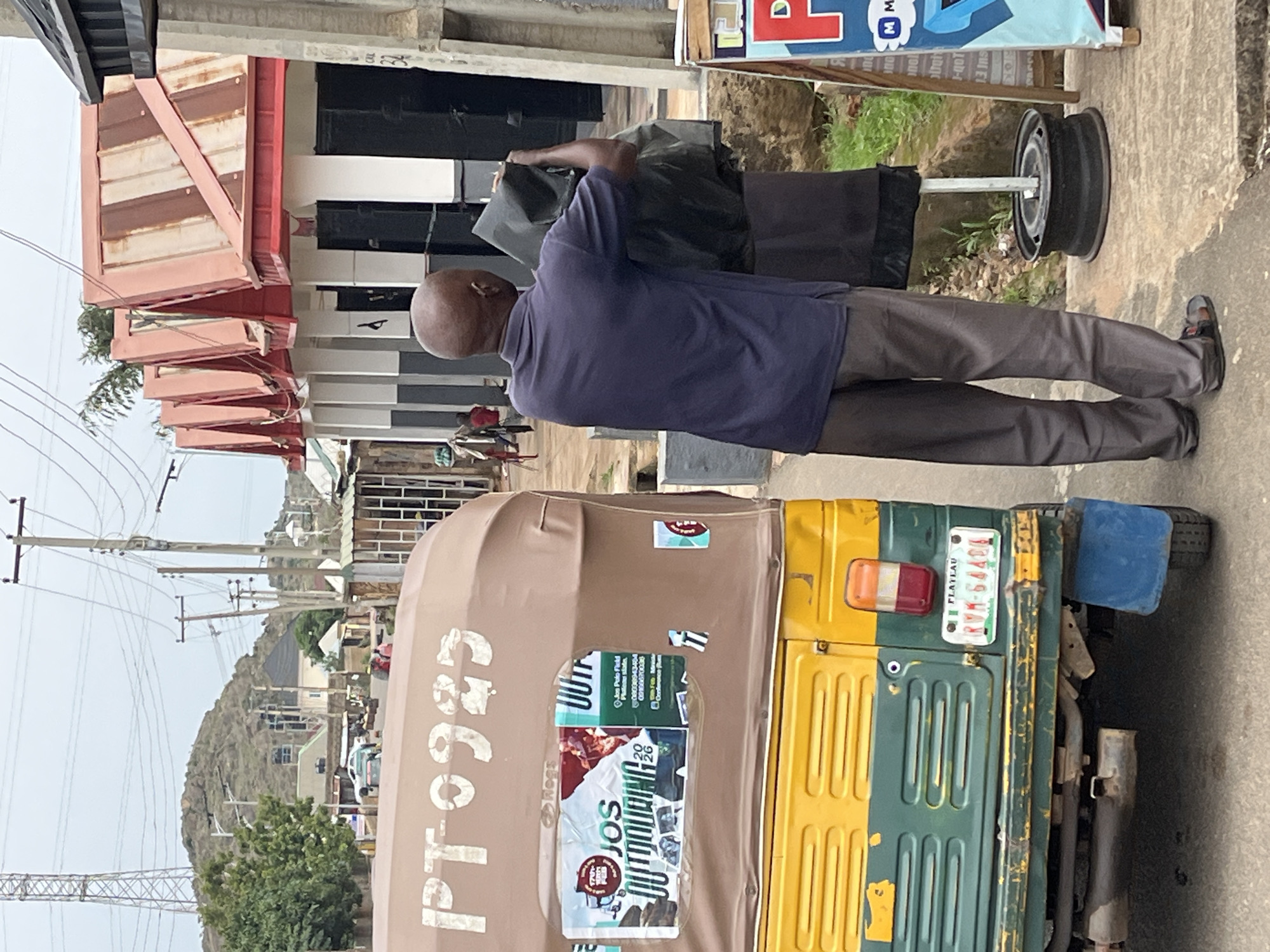A street scene in Barakin Akawo, Nigeria, features a man wearing a navy shirt and gray trousers. He stands beside a commercial vehicle (likely a motorized tricycle) that has the name "JOS" visible in a sticker on the side, along with the license plate "RVK-64406" and the number "PT0925". The vehicle has a predominantly green and yellow color scheme. A pedestrian is present in the background. Buildings with red and white awnings are on the left. Utility poles with power lines are visible against a hilly background.