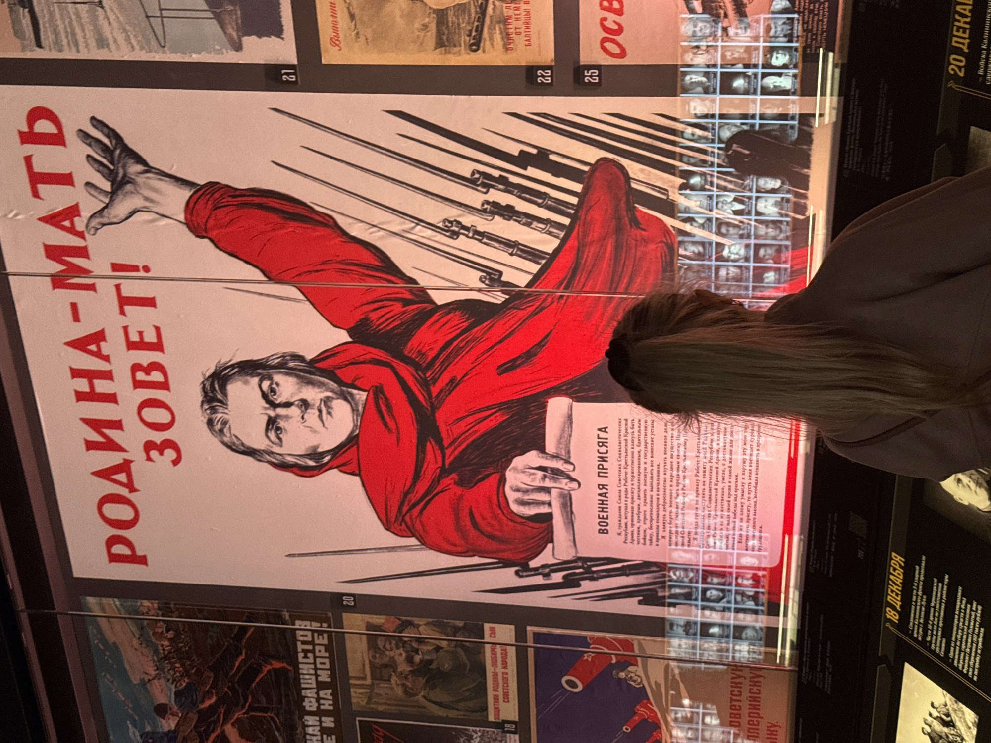 The image shows a museum exhibit featuring World War II propaganda posters in Zelenaya Roshcha, Russia. In the foreground, a young woman with blonde hair pulled back in a ponytail, wearing a brown hooded jacket, is viewed from behind as she looks at a large poster. The poster, predominantly red and white, features a woman's face with a stern expression and a red scarf around her neck, her right arm raised with an open palm, and several rifles pointed upwards behind her. Large red Cyrillic text above the woman reads "РОДИНА-МАТЬ ЗОВЕТ!" (Motherland Calls!). Below the woman's raised arm, a scroll with the heading "ВОЕННАЯ ПРИСЯГА" (Military Oath) is visible, containing text in Russian.
To the left of this central poster, a smaller poster with the text "НАЙ ФАШИСТОВ! Е И НА МОРЕ!" (Fascists! And at sea!) is partially visible, depicting a scene of soldiers. To the right of the central poster, two other posters are visible: one with a number "21" and a yellow background, and another below it with a number "25" and the text "ОСВ" (likely the start of a word).
Below the main posters, a grid of small portrait photographs is displayed. The overall environment suggests an indoor museum setting with controlled lighting.