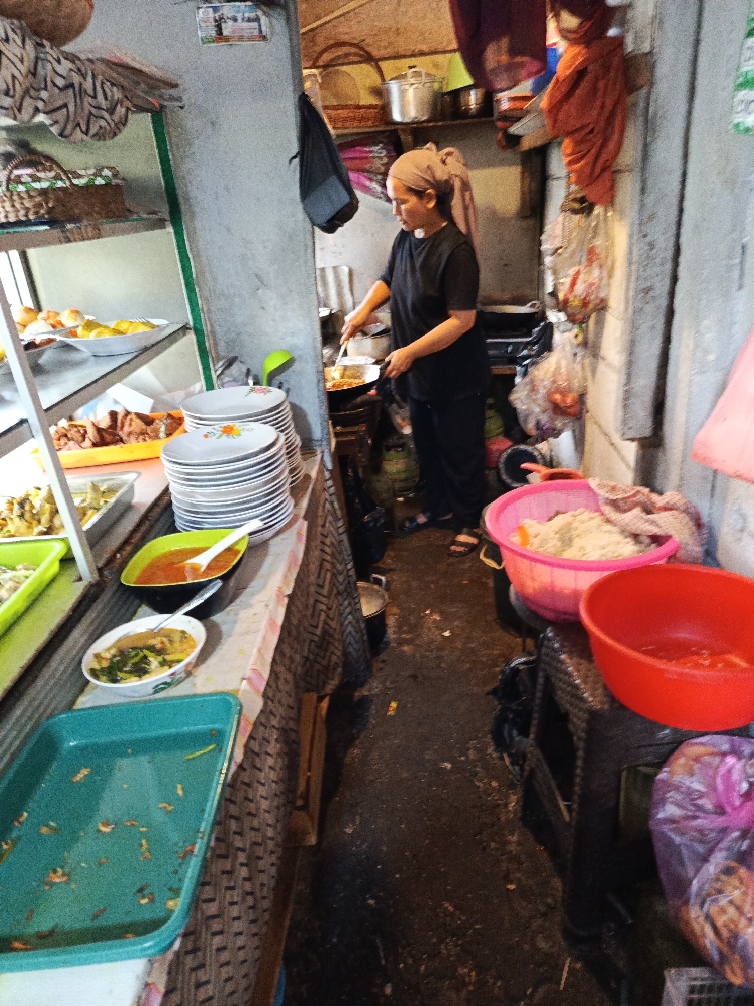 The image depicts a food stall, likely in Tangerang, Indonesia, where a woman in a black shirt and headscarf is cooking. She is standing over a stovetop, stirring food in a pan with a ladle. In the foreground, there are various food dishes and serving bowls, including a large green rectangular tray with some scattered food remnants, a white bowl with a soup-like dish, and a bright orange bowl with a ladle. To the right, there are two large plastic basins, one pink filled with what appears to be rice, and one red, empty. Various items are stored on shelves and hanging on the wall, including baskets, bags, and kitchen utensils. The environment appears to be an outdoor or semi-outdoor food stall with a concrete floor and a somewhat cluttered but functional setup.