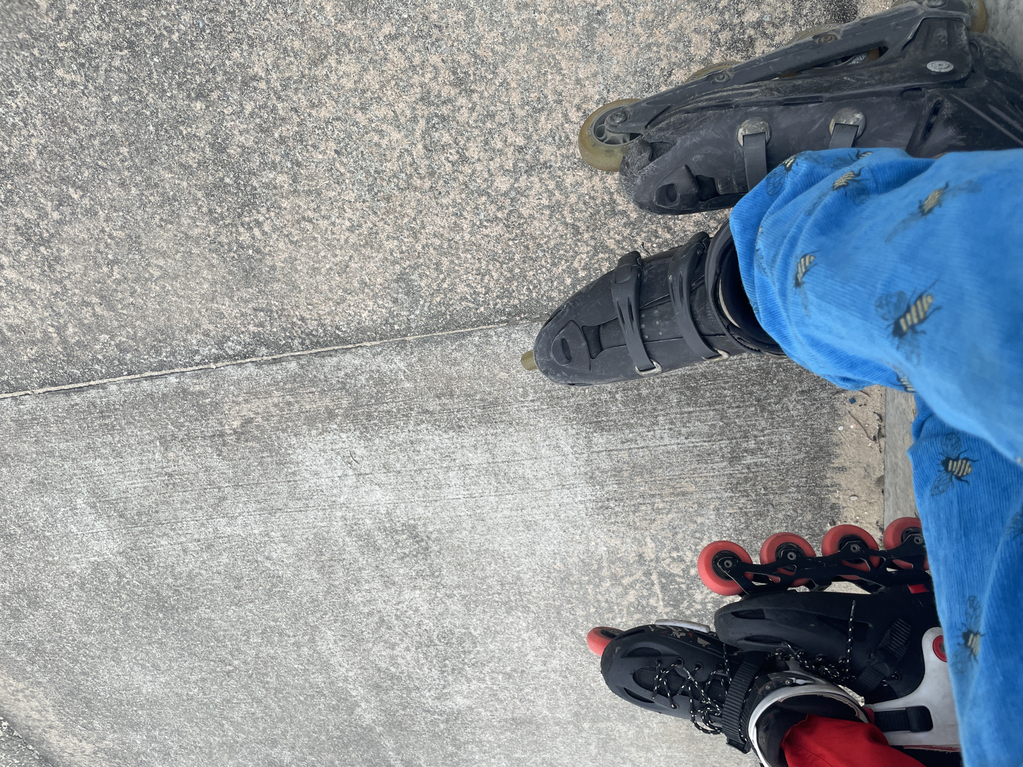 The image shows a close-up, overhead view of two pairs of roller skates on a concrete surface. The roller skates are positioned side-by-side, with the wheels facing upwards on the left and downwards on the right. The concrete surface appears to be a sidewalk or path, with a visible seam running vertically through the center of the image. The left roller skate has black boots with white laces and red wheels. The right roller skate is black with clear or yellowed wheels. Blue fabric with a bee pattern is visible in the lower right quadrant of the image, obscuring part of the right roller skate. The overall environment is outdoors, with the concrete surface suggesting an urban or suburban setting. There are no visible people. The image does not provide any information about the location in the city of Beecroft Point, Nigeria.