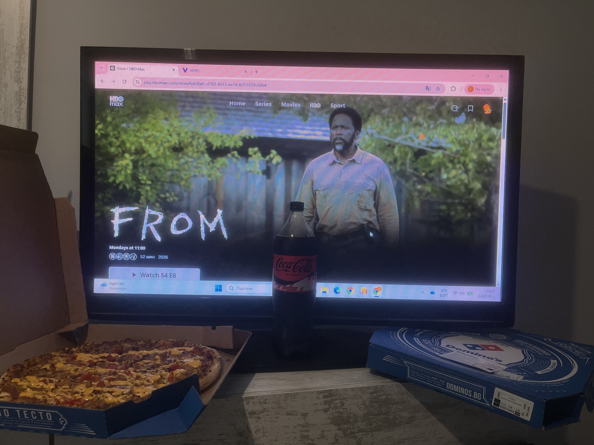 The image displays an indoor leisure setting, likely a living room, in Varna, Bulgaria. A television screen prominently features the HBO Max streaming service, accessed through a Google Chrome browser. The series "FROM" is playing, showing a male character (Harold Perrineau as Boyd Stevens) from the show. On-screen details include "Mondays at 11:00", "52 мин", "2026", and a prompt to "Watch S4 E6". The browser's URL confirms `play.hbomax.com/show/6d6ffafc-d703-4813-ae1d-b253559c60b4`, with another tab for "V VOYO" also open.
In the foreground, an opened Domino's pizza box contains a pizza with toppings that appear to be ground meat, cheese, and diced tomatoes. The box flap reads "O TECTO". A bottle of "Coca-Cola БЕЗ ЗАХАР" (sugar-free) stands between the pizza and the TV. To the right, a closed blue Domino's pizza box displays "DOMINOS.BG" and a label with details like "Carry out Planet Mel" and a contact number "070014444".
The computer's taskbar is visible at the bottom of the screen, indicating a Windows operating system set to Bulgarian ("БГР" for language, "Търсене" for search, "На пауза" for video status). The system clock shows the time as "15:37" and the date as "2.6.2026 г.". A weather notification states "Light rain Tomorrow". The dim lighting of the room, despite the afternoon time displayed on the clock, suggests a cozy atmosphere for watching a show while enjoying pizza and a beverage.