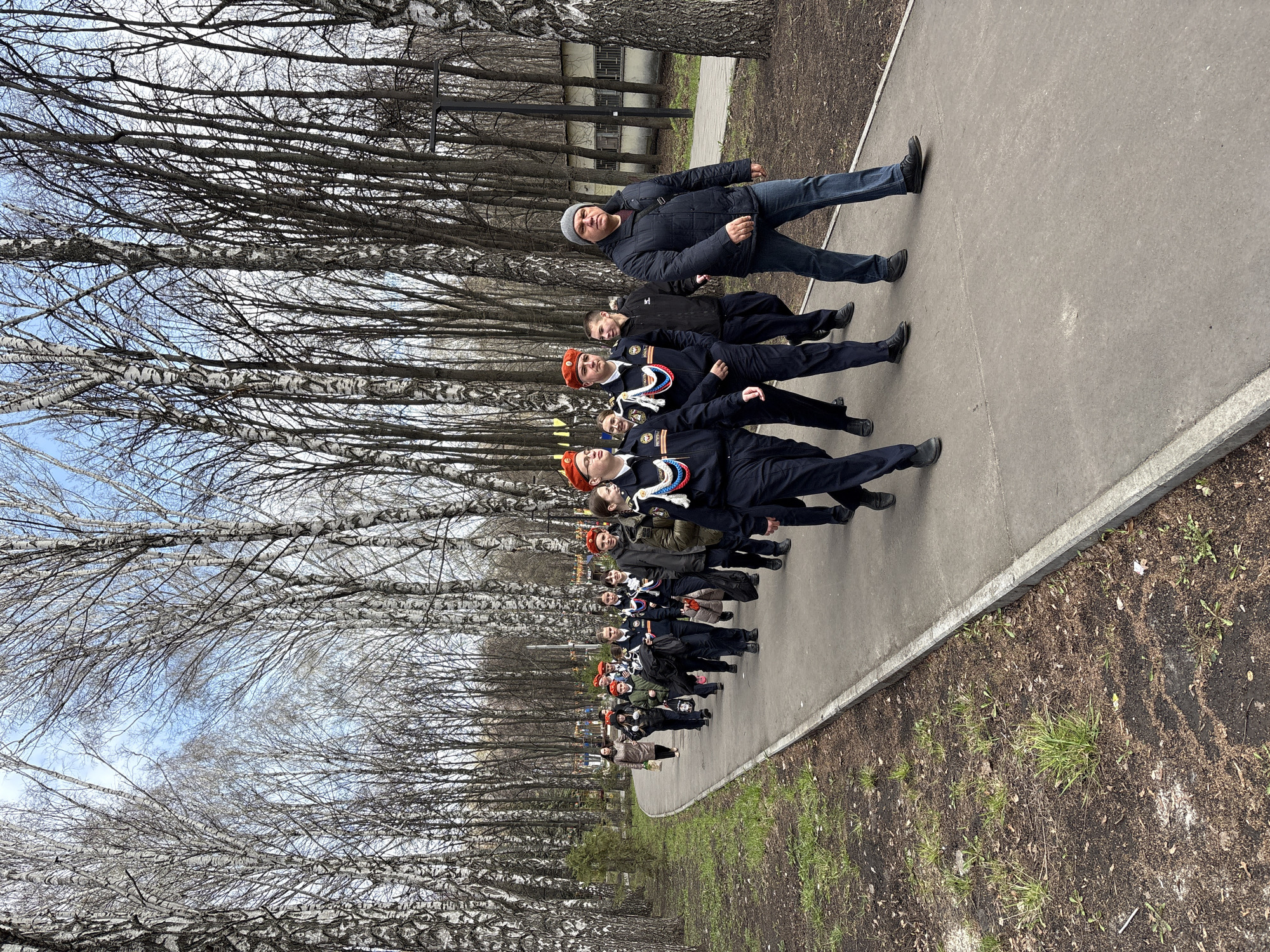 A group of people stand on a paved path in what appears to be a city park in Ulyanovsk, Russia. The group includes uniformed students and adults, possibly instructors or chaperones. They are arranged in a line. Trees, many birch trees, line the path. A building is visible in the background. The ground beside the path shows signs of early spring.