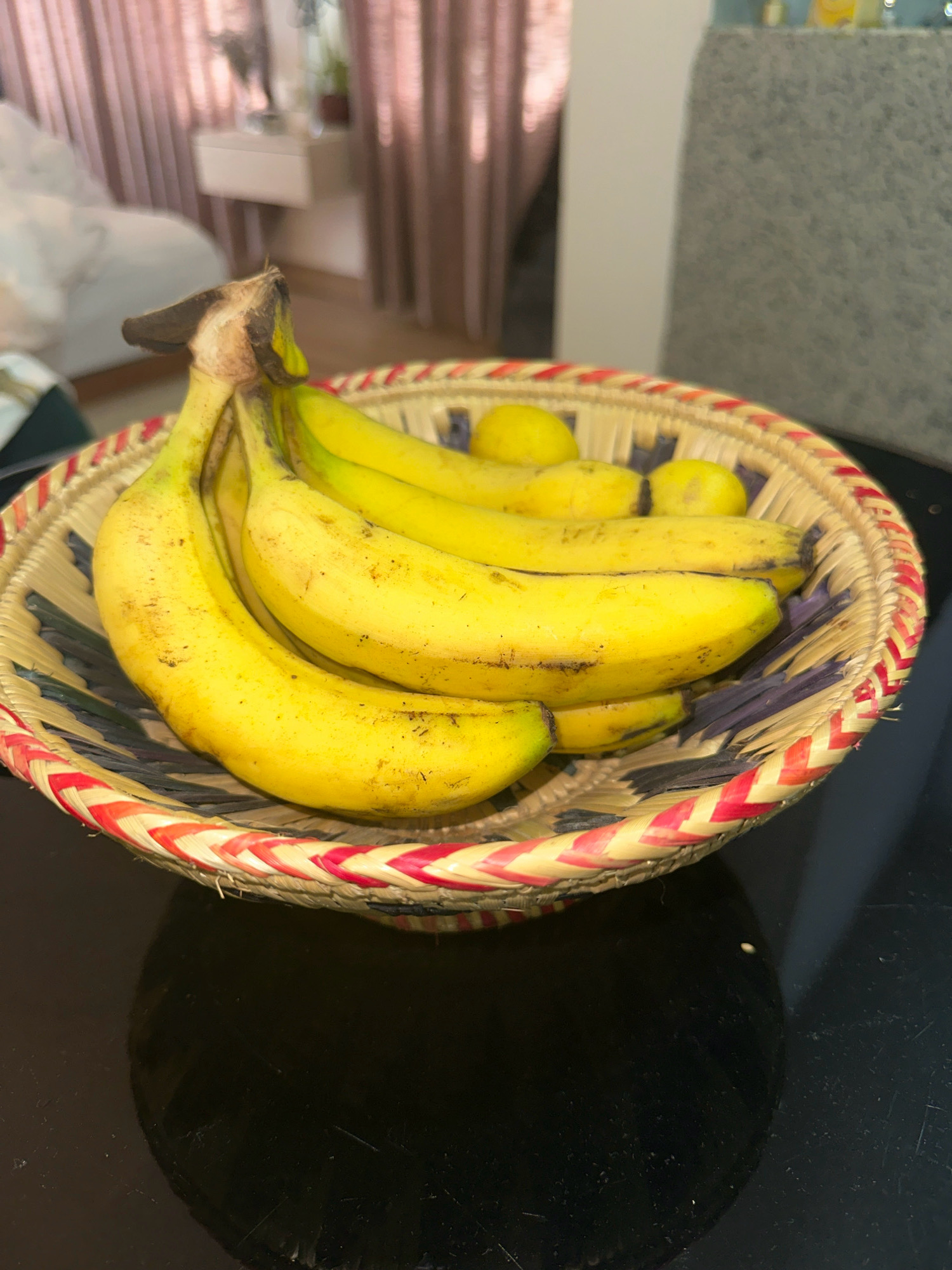 A woven basket containing a bunch of ripe bananas and a few lemons sits on a reflective black surface. The basket is made of straw and decorated with red and yellow braided trim and dark purple woven strips. The bananas are a vibrant yellow with some brown speckles, and one banana at the front has a slightly bruised stem. The lemons are small and bright yellow. In the background, soft pink curtains are visible, along with furniture that suggests an indoor setting, possibly a bedroom. The overall scene is well-lit, with the focus on the fruit in the basket.