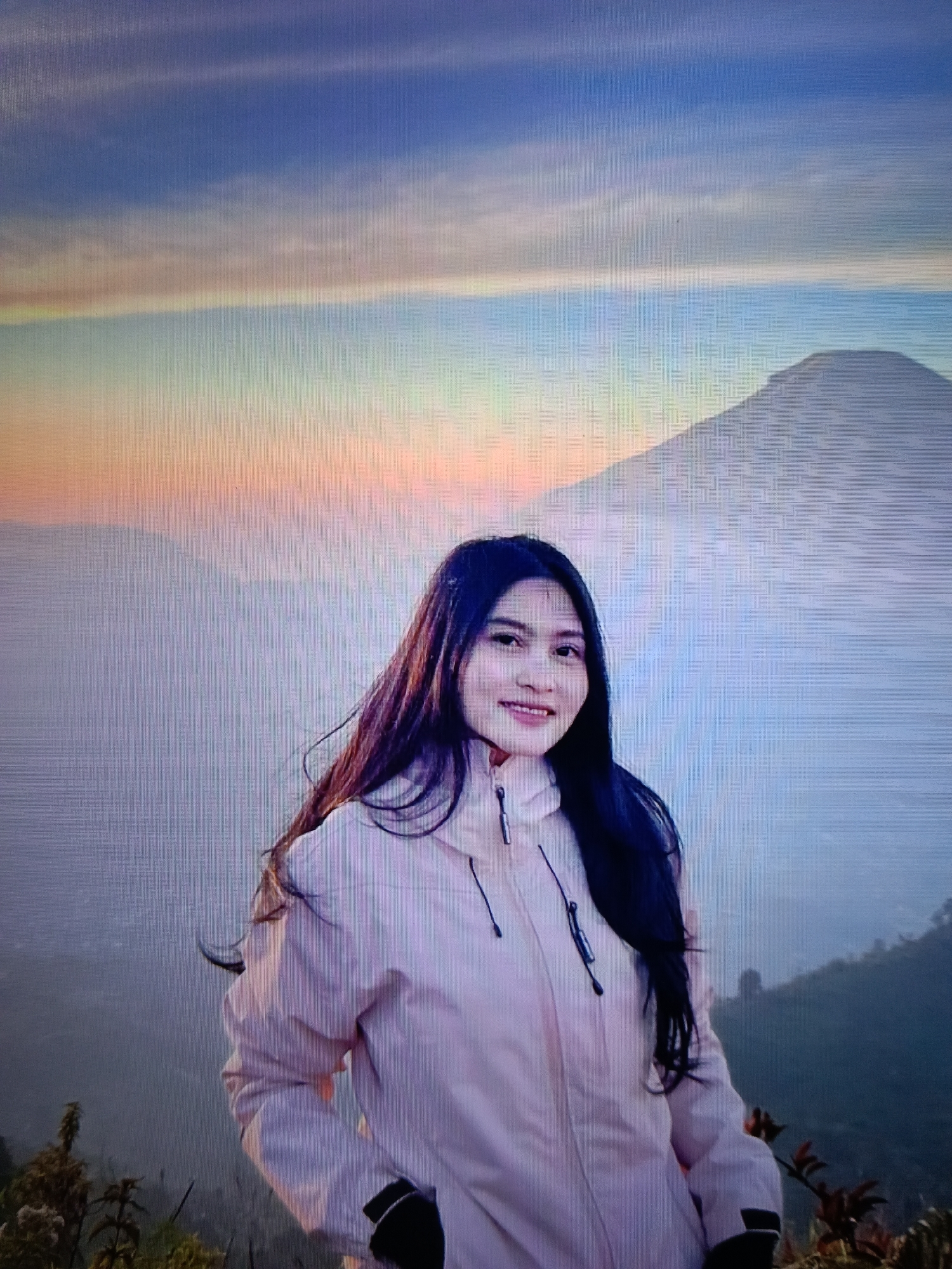 A young woman with long, dark hair is posing for a photo outdoors, with a mountain range and a colorful sky in the background. She is wearing a light pink jacket with black zippers and black cuffs on the sleeves. The sky is a gradient of blue, yellow, and pink, suggesting either sunrise or sunset. The mountain behind her appears to be covered in mist. The foreground shows some vegetation. The image is taken from a slightly low angle, looking up at the woman. The scene does not depict any specific landmarks or elements that would identify it as being in the city of Raha, Indonesia.