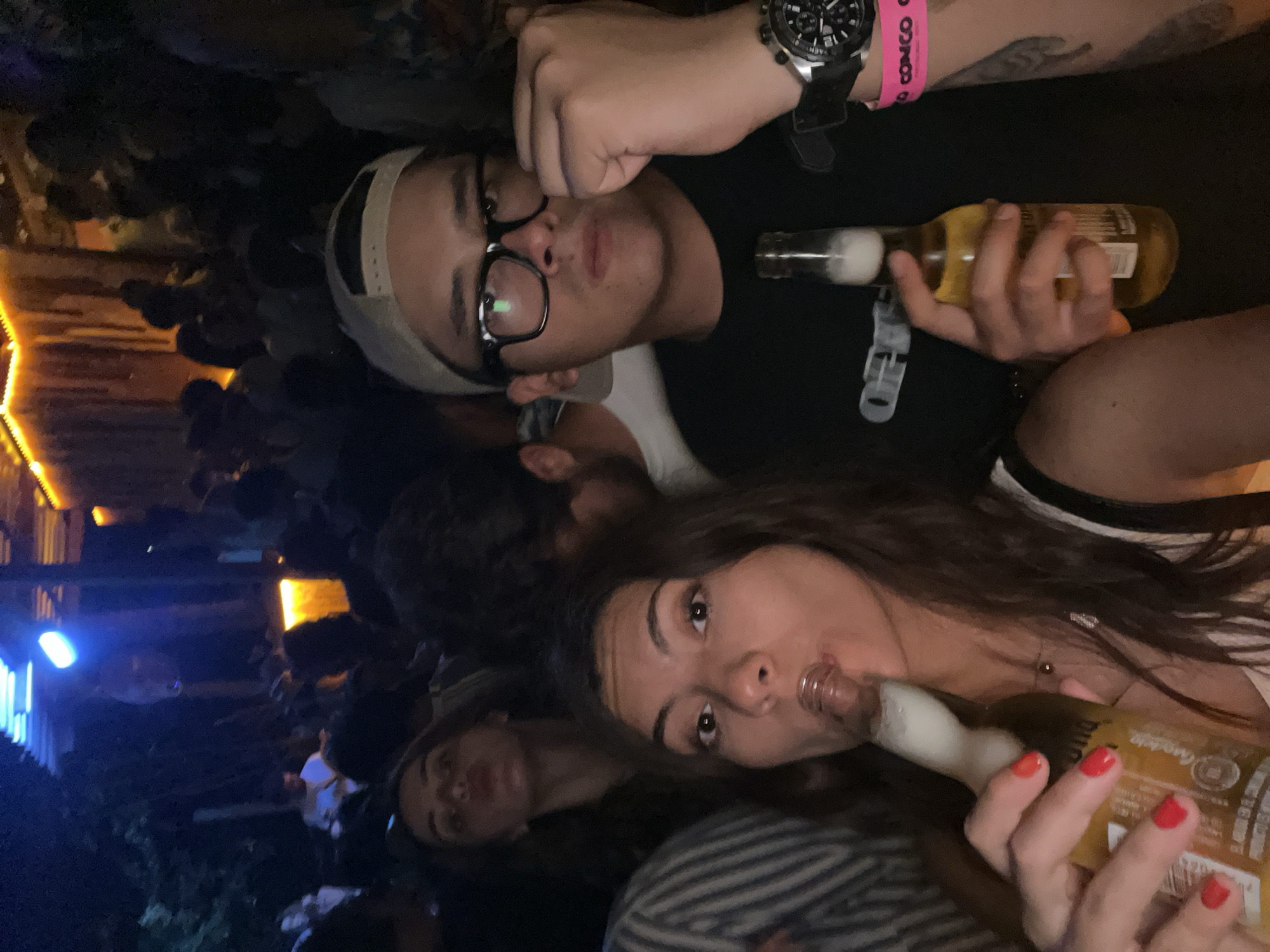 Two young adults pose for a selfie while drinking beer at night in what appears to be a crowded outdoor venue. A third person is partially visible in the background, also appearing to be drinking. The man wears glasses and a baseball cap, while the woman has long hair and red nail polish. They are holding Corona beer bottles. The environment is dimly lit with some lights visible in the background and the surroundings suggest that this venue is in Puerto Escondido, Mexico.