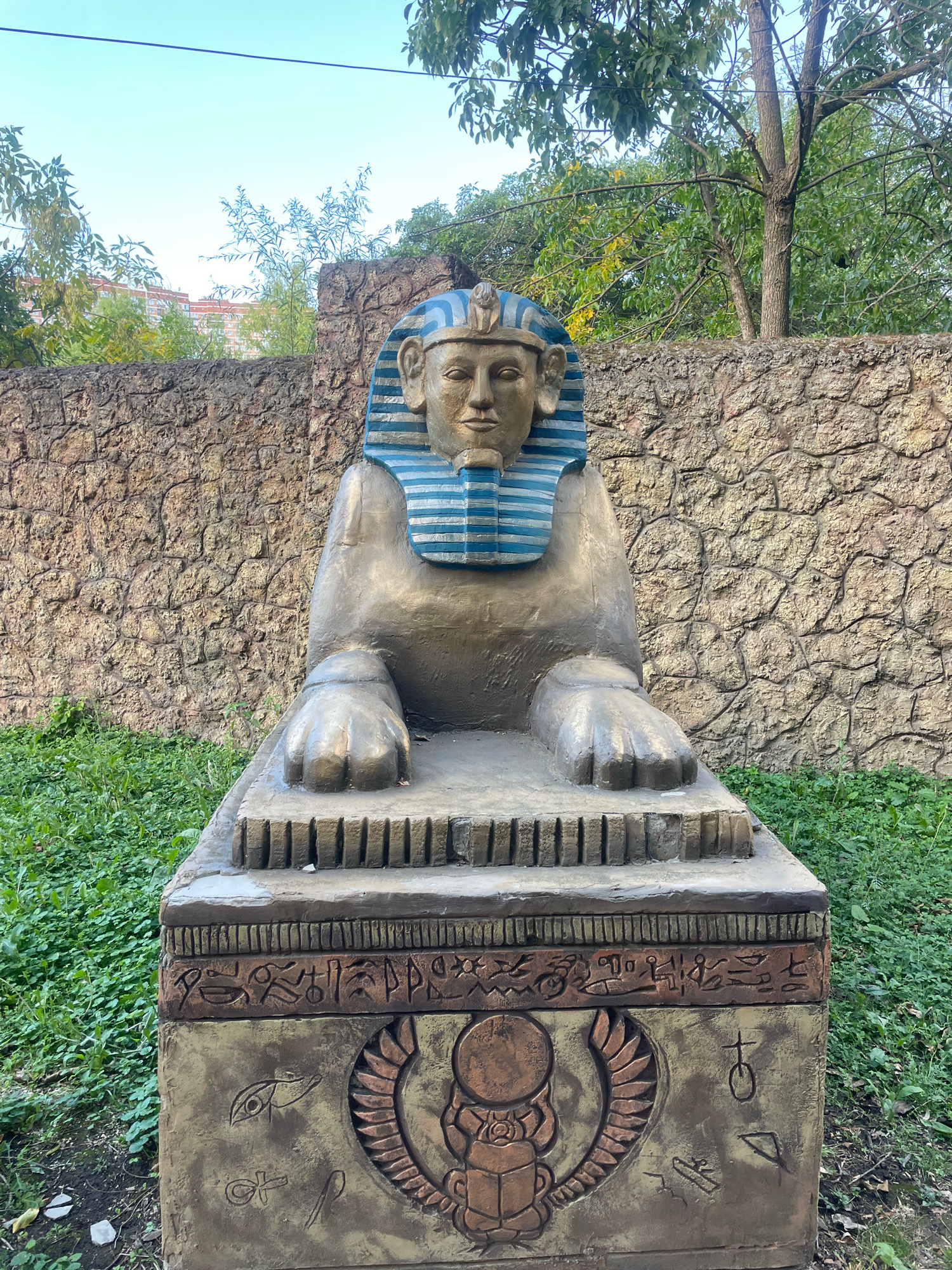The image shows a statue of a Sphinx in an outdoor setting, likely a park or garden. The Sphinx is a golden-brown color with a blue and gold striped headdress. It is seated on a stone pedestal that is decorated with Egyptian hieroglyphs and a scarab beetle motif. The background consists of a rough, stone wall and lush green grass. Trees with green leaves are visible behind the wall, and a clear blue sky is overhead. The statue appears to be a replica or a decorative piece rather than an ancient artifact. The overall scene suggests a publicly accessible area with a theme related to ancient Egypt. Based on available information, this Sphinx statue is located in the Park of Culture and Recreation in the city of Krasnodar, Russia.