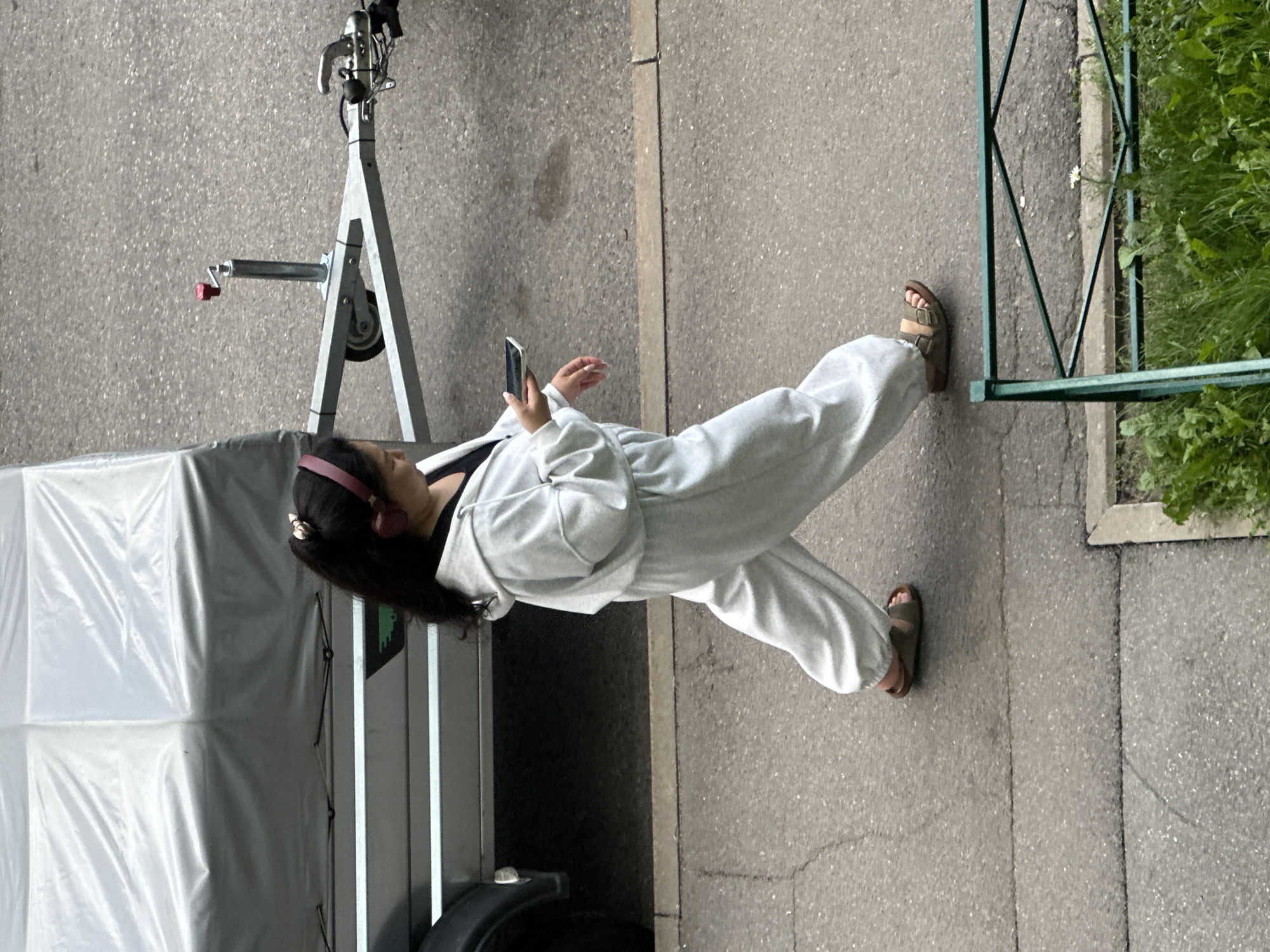 In Murino, Russia, a young woman with dark hair tied in a ponytail and wearing pink headphones is walking on a paved sidewalk next to a gray trailer. She is dressed in a light gray tracksuit, including a hooded jacket and loose-fitting pants, and wears brown sandals. The woman is holding a white smartphone in her right hand and appears to be looking at it. The trailer is a light gray color with a white cover. The environment is urban, with a textured asphalt ground and some greenery in the foreground, suggesting a residential or commercial area.