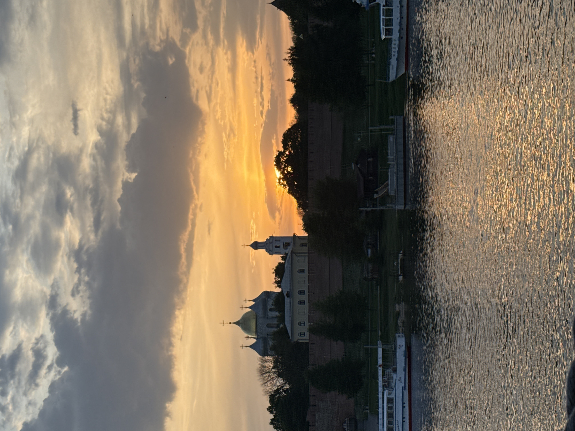 The image depicts a sunset scene in Veliky Novgorod, Russia. The sky is filled with a mix of grey and yellow clouds, with the sun setting behind the treeline, casting a golden glow. The outline of the Saint Sophia Cathedral is visible, along with its dome and crosses. In the foreground, a river runs vertically, reflecting the golden hues of the sunset, with light reflecting. A boat is at the edge of the river. The fortress wall is visible, along with some trees.