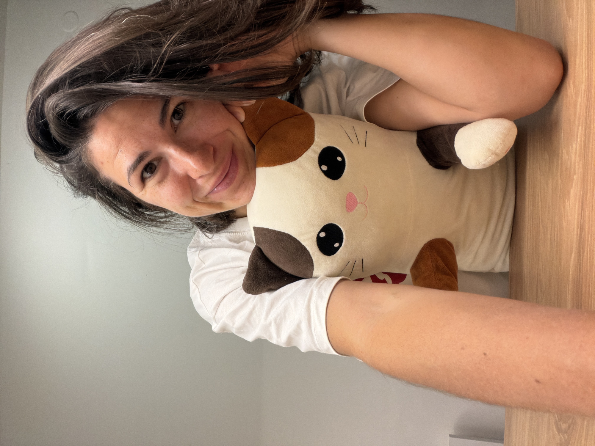 A woman with dark hair is holding a plush cat toy. She is wearing a white t-shirt. The background is a light gray wall and a wooden surface. The location cannot be identified as Budva, Montenegro from the image.