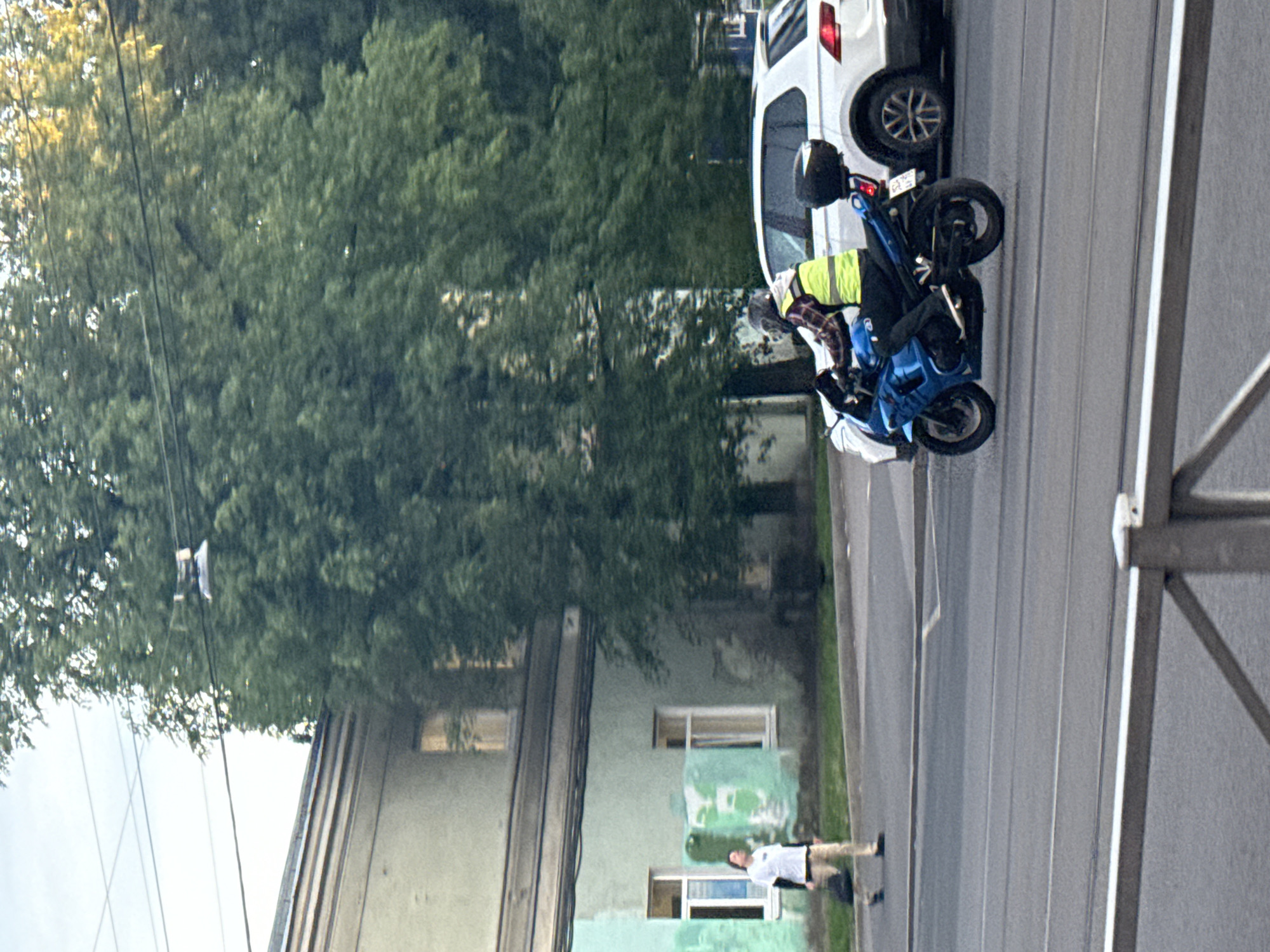 A motorcyclist wearing a helmet and a bright yellow vest rides a blue sportbike on a city street in Saint Petersburg, Russia. The motorcyclist is seen from the side, with their left leg extended forward. Behind the motorcycle, a white SUV is partially visible. In the background, a light green building with windows and overhead wires can be seen. To the left of the building, a pedestrian in a white shirt and beige pants walks across the street, carrying a dark bag. The street has visible tram tracks. The environment appears to be an urban setting with a tree providing shade on the right side of the frame.