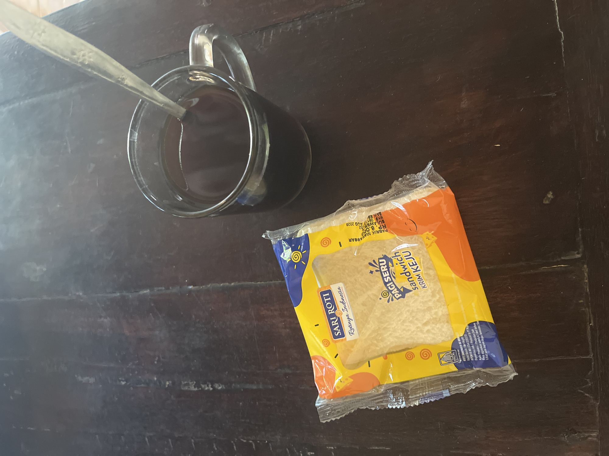 In Bumi Mulyo, Indonesia, a glass mug filled with dark liquid, presumably coffee, rests on a wooden table. A silver spoon is partially submerged in the mug. Next to the mug lies a packaged sandwich, its yellow and orange wrapper displaying the "SARI ROTI" and "PAGI SERU" brands, indicating it is a cheese cream sandwich. The table's surface shows a dark, weathered wood grain.