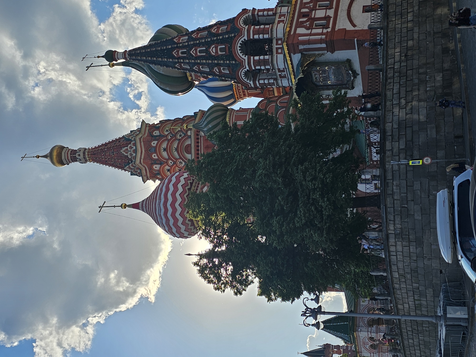 The image displays a vibrant architectural landmark, strongly resembling St. Basil's Cathedral, under a bright, partly cloudy sky. The photograph is oriented vertically, showing the towering, colorful domes and spires reaching towards the sky.
The central subject is a multi-domed building featuring intricate red brickwork, white trim, and decorative patterns. Several onion domes are visible, including one prominently striped in red and white chevron patterns, another in solid green, and another in blue and white stripes. Golden finials and crosses adorn the peaks of the domes and spires. A large, dense green tree occupies the middle ground, its foliage partially obscuring the lower sections of the cathedral but backlit by the sun, creating a silhouette effect.
Below the main structure, a paved area, possibly a street or square, is visible with a cobblestone-like pattern. A black metal railing separates the building's immediate vicinity from this paved space. Several small figures of people can be seen on the ground level, walking or standing, suggesting public access or a tourist area. On the far right, parts of parked vehicles, including a white car-top carrier and a dark car, are visible. A black ornate streetlamp with multiple arms stands in the lower right foreground.
The sky is a mix of bright blue patches and large, fluffy white and grey clouds. The lighting suggests it is daytime, with the sun positioned to create some backlighting and shadows. A notable detail is a yellow road sign on the paved area, which clearly shows a white bicycle symbol with a black rider and a black arrow pointing to the right, indicating a cycling route or warning.