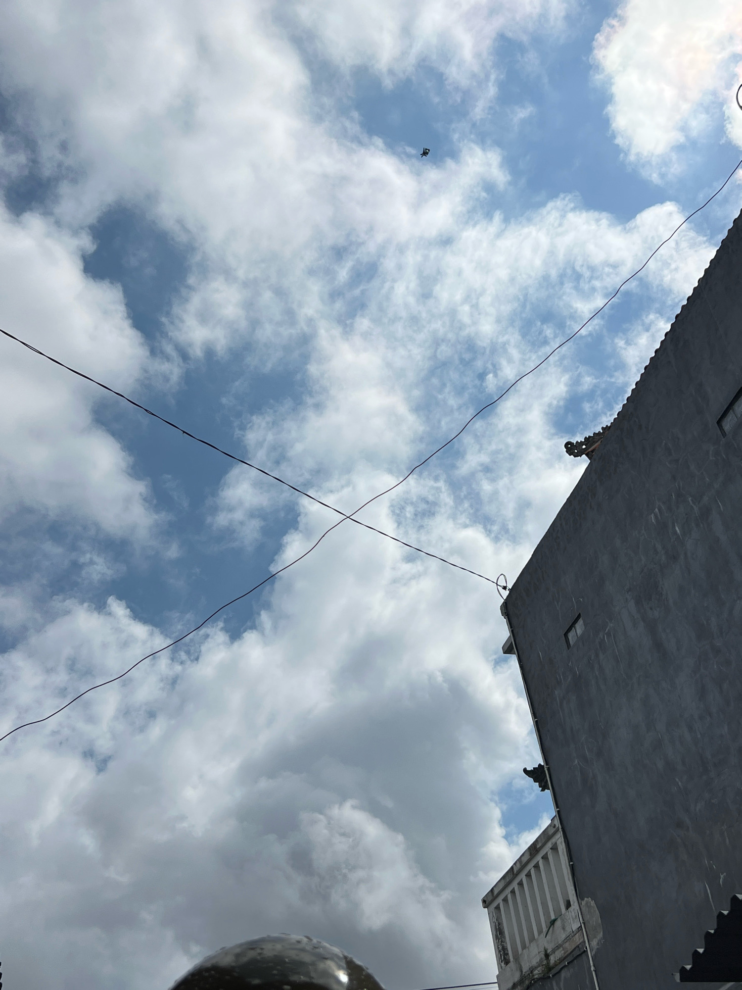 The image captures a view of the sky over a cityscape, with a prominent building on the right. The sky is partly cloudy with patches of blue visible. Two dark power lines diagonally traverse the frame, intersecting in the upper half. On the right side of the image, a grey building rises, its angular facade partially obscuring the sky. The roofline of the building features decorative elements, including what appears to be a gargoyle-like figure. Some windows are visible on the building's exterior. The lower portion of the image shows a glimpse of what might be a reflective surface, possibly part of a vehicle or another object. A tiny, dark object, possibly a drone or a bird, is visible in the sky in the upper left quadrant. The lighting suggests it is daytime. The context of Denpasar, Indonesia, implies this is likely an urban or suburban environment.