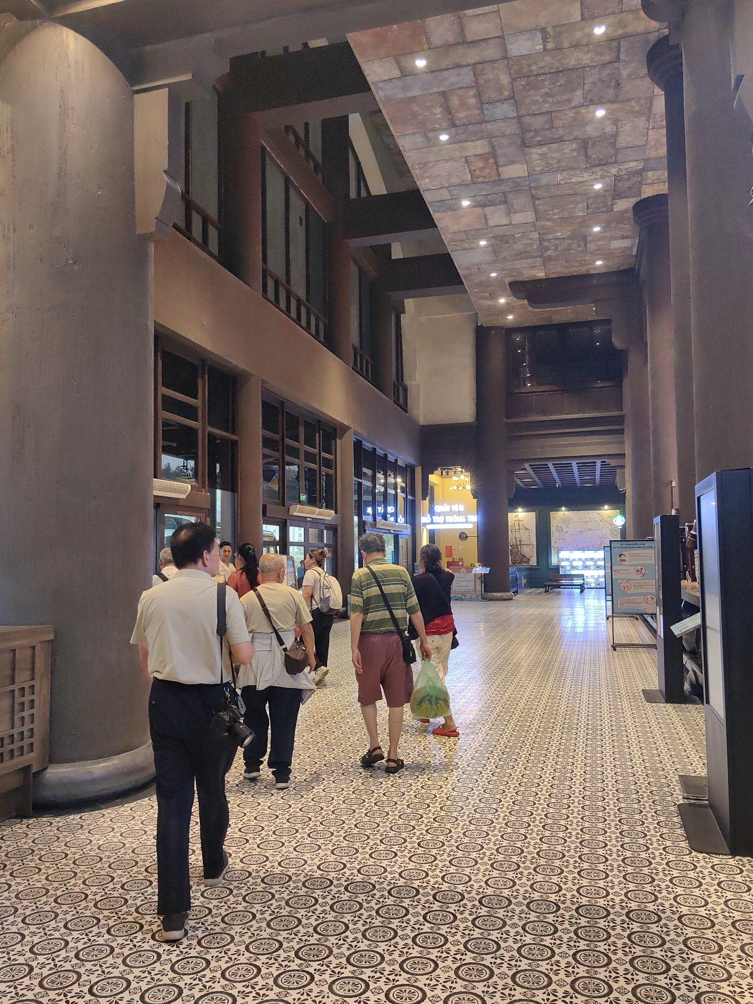 A group of people walks through a well-lit interior space with tiled floors and large pillars. The architecture features a mix of modern and traditional elements with exposed wooden beams and high ceilings. Some people appear to be entering or exiting a doorway, while others are further inside the space. The location may be in Bai Chay Ward, Vietnam, though this is unconfirmed.