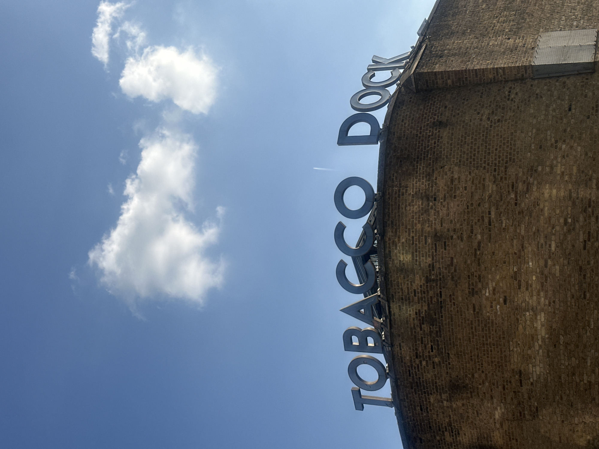 The image displays the exterior of a large, robust brick building under a clear blue sky with scattered white clouds. The photograph is taken from a low angle, looking upwards at the building's facade, and is oriented vertically, rotated 90 degrees counter-clockwise.
Prominently featured on the building's upper edge are large, silver-colored metallic letters spelling out "TOBACCO DOCK". The letters are three-dimensional and stand out against the brickwork. The building itself is constructed from dark, weathered bricks, suggesting historical significance and durability.
The scene depicts a sunny daytime, indicated by the bright blue sky and the illumination on the building and clouds. A faint, thin white line, likely an airplane contrail, is visible streaking across the sky. There is no specific activity or event taking place, as the image is a static shot of the building's exterior.
Based on the visible text "TOBACCO DOCK" and the provided context, the location is Tobacco Dock in London, United Kingdom. The architecture is consistent with historic dockside buildings found in the area.
