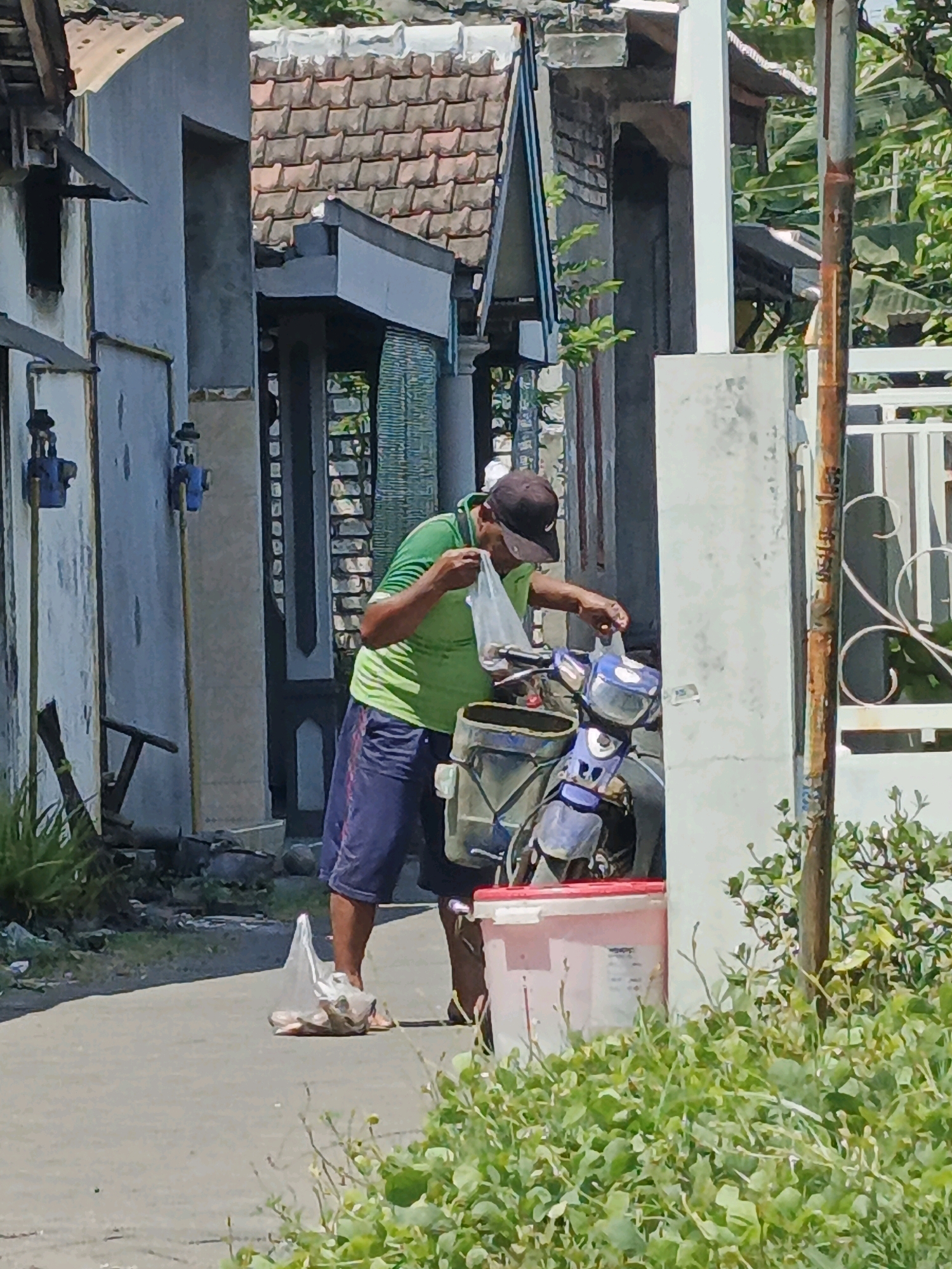 In Ketegan, Indonesia, a man in a green t-shirt and blue shorts is standing next to a blue motorcycle on a paved pathway. He is holding a clear plastic bag filled with items. The motorcycle has a basket attached to the front, and a pink and white bucket is situated next to it on the ground. The man appears to be in the process of placing the bag into the basket. The environment suggests a sunny day with buildings and vegetation visible in the background. The pathway is lined with grass and bushes on the right side of the frame.
