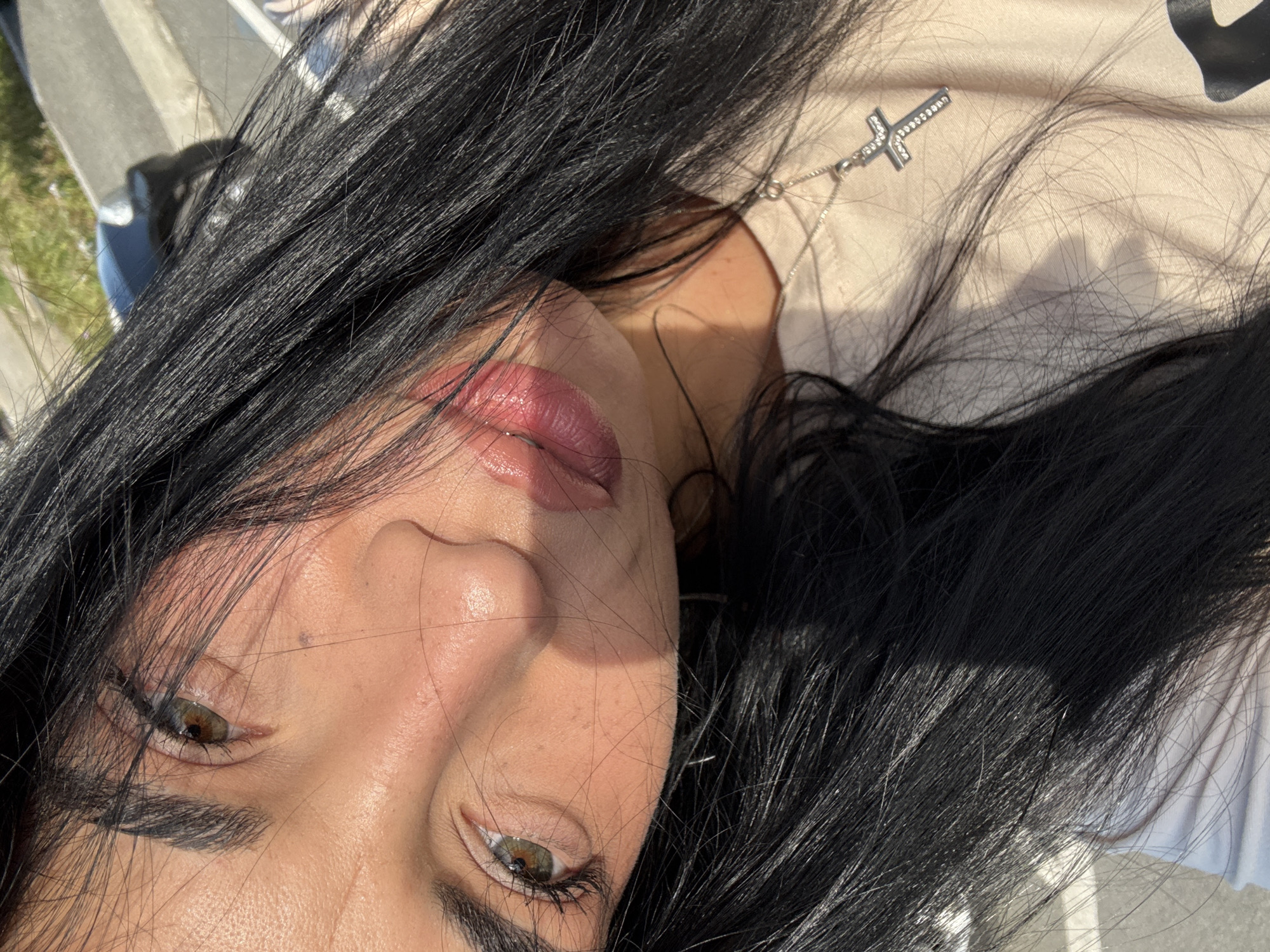The image shows a close-up, angled view of a person's face. The individual has dark hair, fair skin, and hazel-colored eyes. A silver cross pendant necklace is visible. The person is wearing light pink lip gloss. The background is out of focus, but suggests an outdoor setting. The location is not discernible as Nesebar, Bulgaria based on the given image.