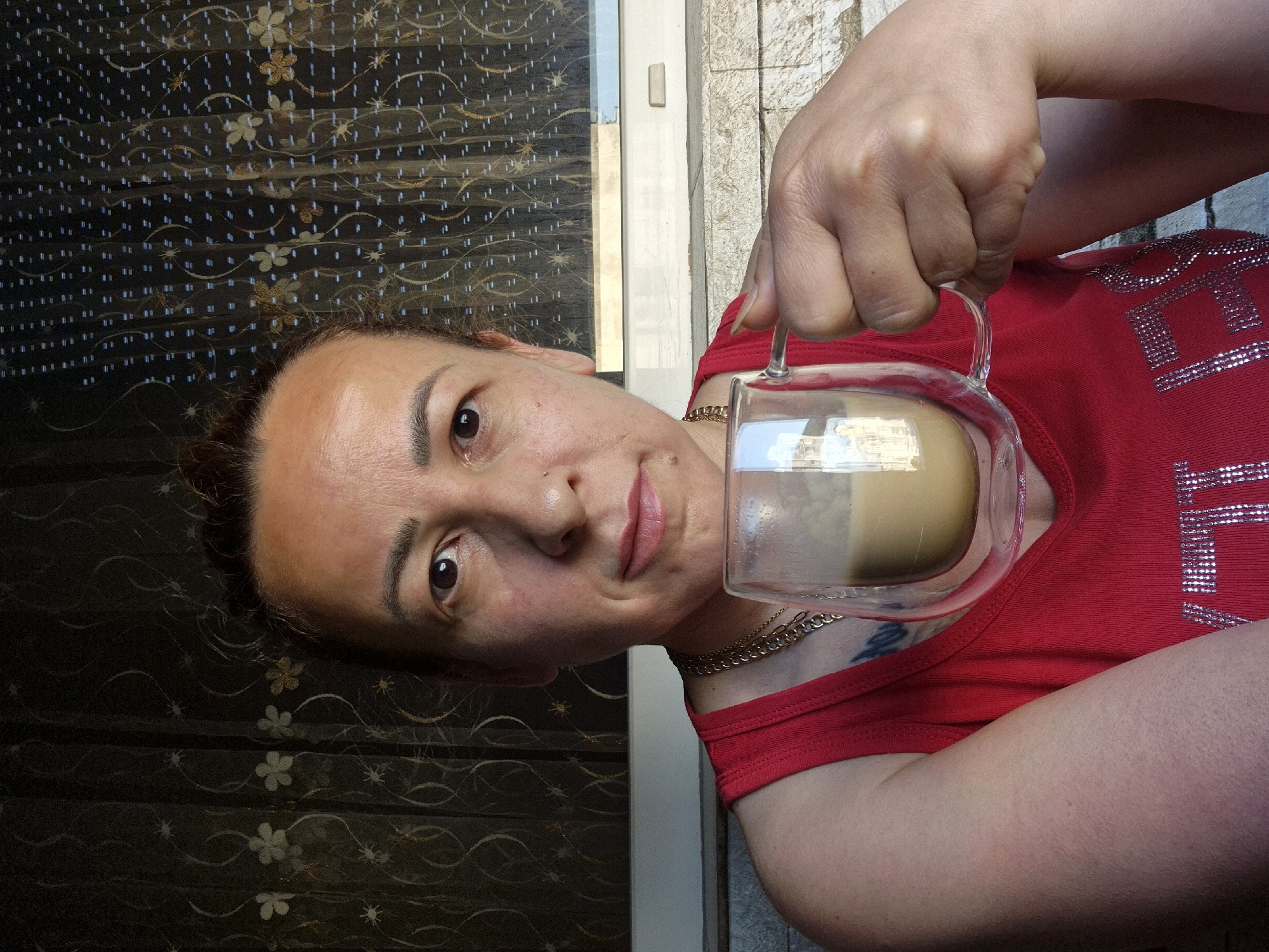The media file captures a portrait of a woman indoors, holding a clear glass mug with a light brown beverage, likely coffee or latte. She is positioned slightly off-center to the left, looking directly at the camera with a neutral expression. Her dark hair is pulled back, revealing her face which has several small moles and a small stud nose piercing on her left nostril. She wears a bright red sleeveless top with sparkly silver elements forming what appears to be part of a word or design, with "VE" being partially visible. A gold chain necklace is visible around her neck, and a tattoo can be seen on her left shoulder/upper arm area, peeking out from under her top.
The background on the left side of the image features a dark, patterned fabric, possibly a curtain or blind, adorned with light-colored floral or star-like motifs and small blue/white dash patterns. On the right side, behind her right shoulder and arm, there's a lighter, more structured background, resembling a white door frame or window frame against a light-colored wall or stone facade, suggesting an indoor setting with natural light source nearby. The mug she holds is a double-walled glass design, showcasing the layers of the drink. The activity is a simple pose for the camera while holding a drink. The lighting suggests it is daytime.