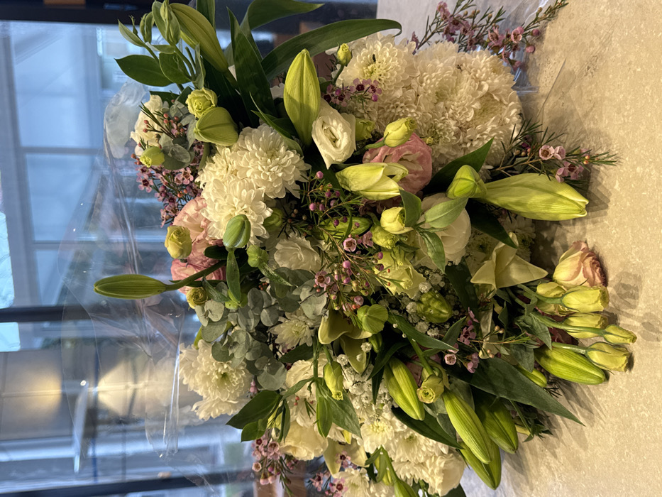 A large, mixed floral bouquet is displayed on a light beige, textured countertop indoors, located in Tel-Aviv, Israel. The bouquet is primarily composed of white chrysanthemum poms, numerous closed green lily buds, light pink and cream lisianthus flowers and buds, small purple-pink waxflowers, and green eucalyptus leaves, interspersed with sprigs of white gypsophila. It is partially enveloped in clear cellophane wrapping. The background reveals a large window reflecting interior light fixtures and an ambiguous exterior view. There are no individuals, explicit actions, or interactions depicted within the frame.