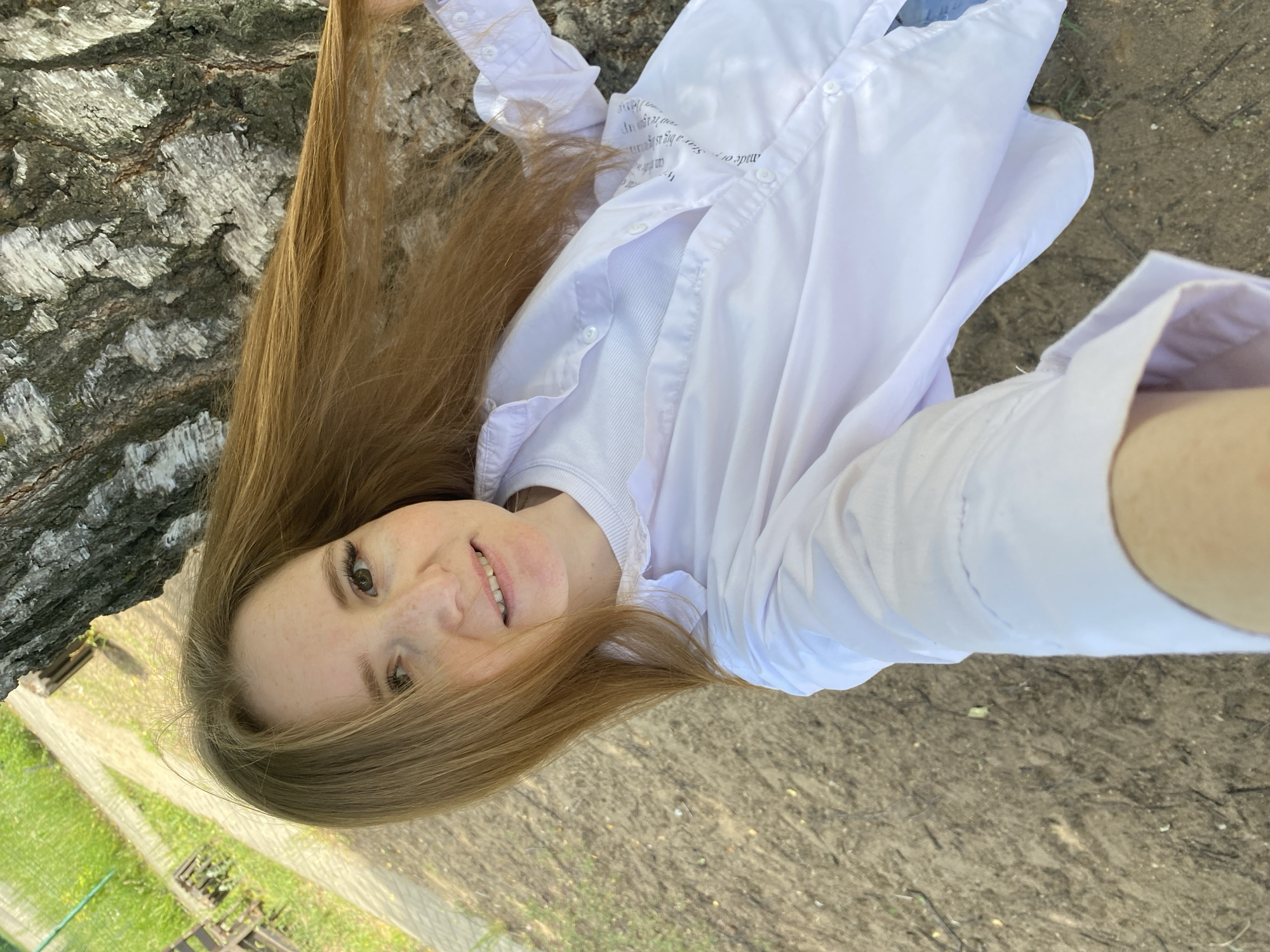 A young Caucasian woman with long, reddish-brown hair and freckles smiles at the camera, holding her hair back with her right hand. She is wearing a white collared shirt over a white t-shirt. The woman is standing outdoors next to a large tree trunk with rough, textured bark. The ground is a mix of dirt and small pebbles, with some dried leaves. To the left of the woman, a paved path is visible, bordered by grass and some wooden structures. The scene appears to be in a park or a natural area, possibly within the city of Chaikovsky, Russia, given the context. The lighting suggests it is daytime.