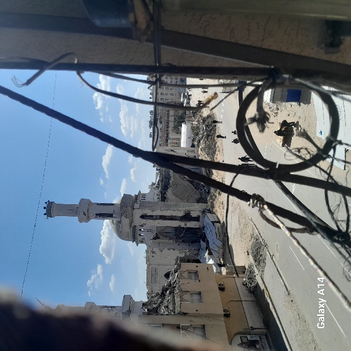 This image captures a wide view of a street scene in Khan Yunis, Palestinian Territory, under a clear blue sky with scattered white clouds. The primary subject is a large, multi-domed mosque with two prominent minarets, extensively damaged around its main dome and lower sections, revealing internal structures and exposed rebar. A multi-story building to the left of the mosque has suffered catastrophic collapse, its upper floors and facade reduced to a large pile of rubble on the ground.
The asphalt street, clear of major debris, runs horizontally across the mid-ground, flanked by considerable accumulations of concrete rubble, twisted metal, and shattered building materials from collapsed structures. Approximately 10-15 individuals are dispersed throughout the scene. A small group of 2-3 people are gathered, crouched or standing, over an indistinct object on the ground near a rubble pile on the right side of the street. Other individuals are seen walking along the street.
In the far background, additional urban buildings appear less damaged, and a white tent structure is visible. The immediate foreground is partially obscured by dark, out-of-focus metal bars and wires, indicating the photograph was taken from an elevated position, likely through a window grating or balcony railing. A "Galaxy A14" watermark is present in the lower-left corner of the original image. The scene portrays an urban landscape marked by significant structural damage and ongoing human presence amidst debris.