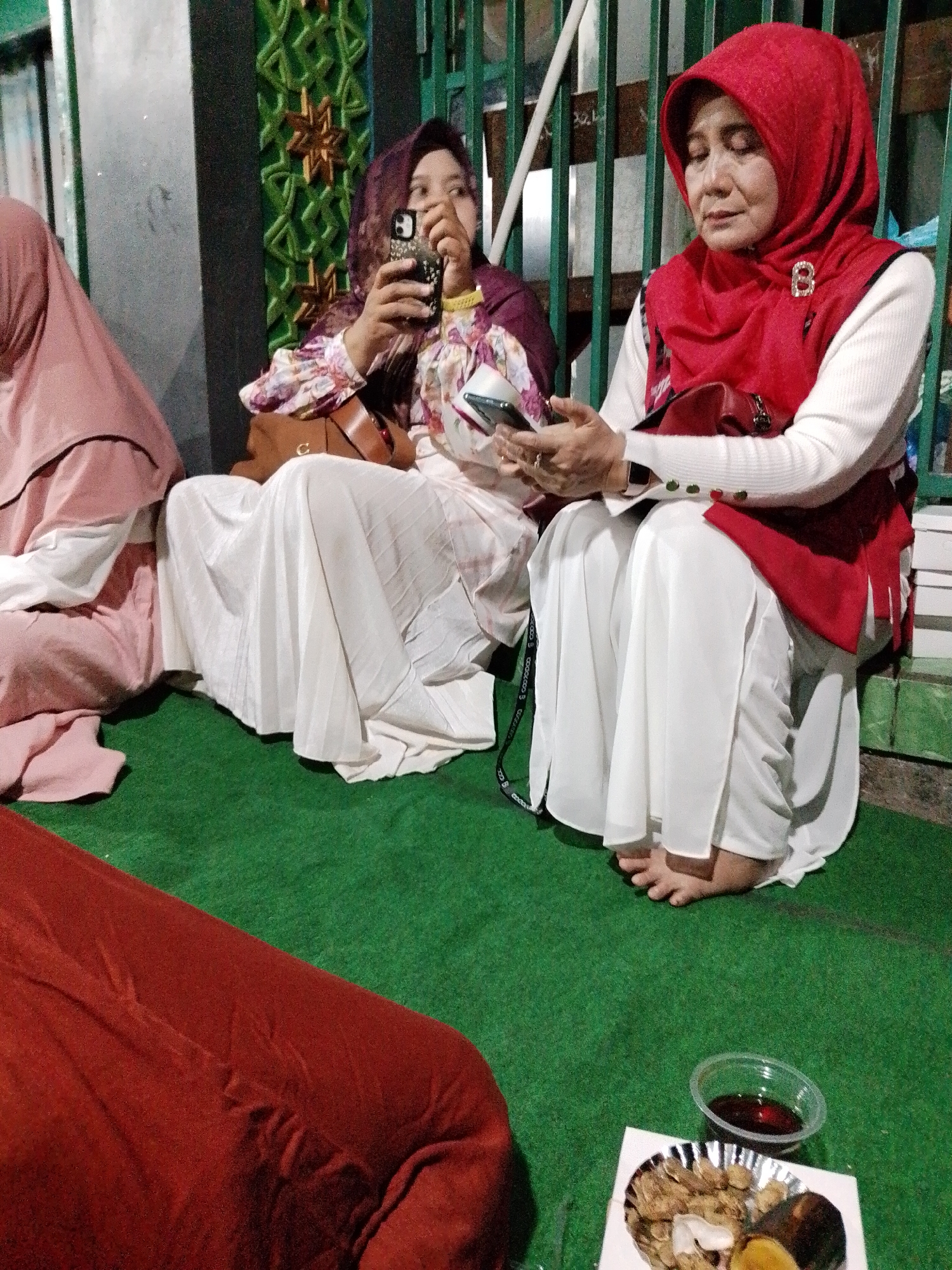 In Kecamatan Pati, Indonesia, a scene unfolds on a green carpeted surface, possibly outdoors or in a covered area with a metallic fence in the background. Three women are present, dressed in traditional Islamic attire.
The woman on the far left wears a pink hijab and a pink outfit. To her right, another woman, also in a hijab (this one a darker purple), is seated and holding a smartphone, actively engaged with it. She wears a patterned blouse with voluminous sleeves.
The woman in the foreground, wearing a bright red hijab, is seated and looking down at her smartphone, which she holds in her hands. She has a white long-sleeved shirt on beneath a red vest, adorned with a decorative brooch. Her white skirt is pleated. Her feet are visible, crossed.
In front of the woman in the red hijab, a small plate with food items, including what appears to be fried snacks and a dark brown piece of food, sits next to a clear plastic container filled with a dark liquid. The green carpet extends across the foreground, with a large red fabric item partially visible at the bottom left. The metallic fence in the background has green painted bars.