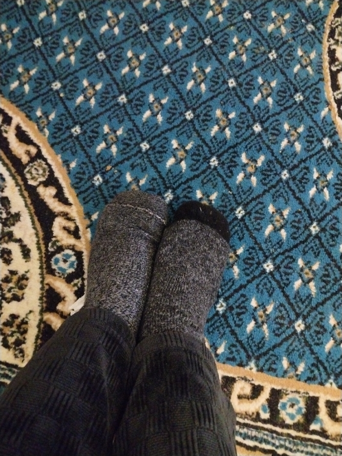 Two feet clad in gray socks rest on a carpet. The socks are different shades of gray, the left foot is a lighter gray, the right foot is a darker gray with a black toe. The person is wearing dark gray ribbed pants. The carpet has a geometric design in shades of blue, beige, and black. A portion of an ornate rug with a circular pattern is visible on the left.
