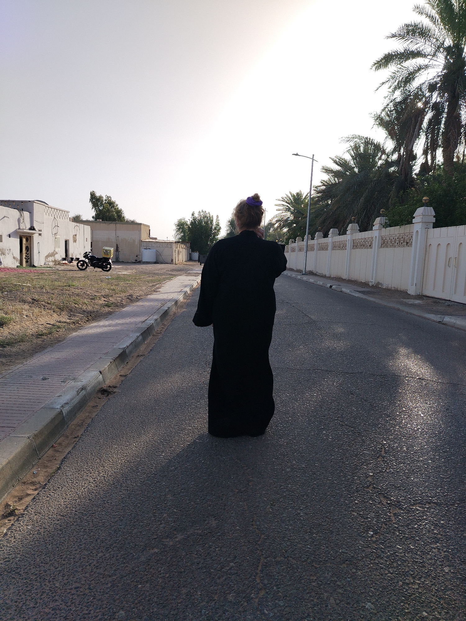 The image presents a residential street scene in what appears to be the city of Al Mas'oudi, United Arab Emirates, characterized by a bright, diffused sunlight. A woman wearing a black robe is walking away from the viewer along the right side of the road. She has a purple hair tie.
To her left is a paved pedestrian path alongside a road with asphalt surfacing and scattered patched sections. A row of white single-story buildings with unique architectural elements is present. A black motorcycle is parked near one of the buildings. A white wall runs along the right side of the road, bordered by tall palm trees.