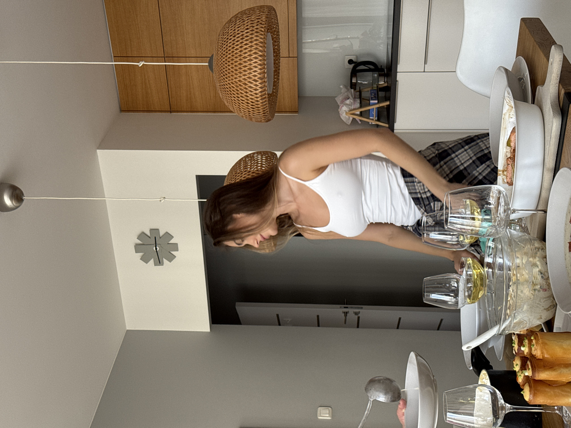 A woman stands at a dining table set with multiple plates and dishes containing food, along with several wine glasses. She is wearing a white tank top and plaid pants. Above the table hangs a pair of decorative wicker lamps. A clock adorns the wall in the background. The setting appears to be an apartment in Warsaw, Poland, based on the architectural style and interior design.