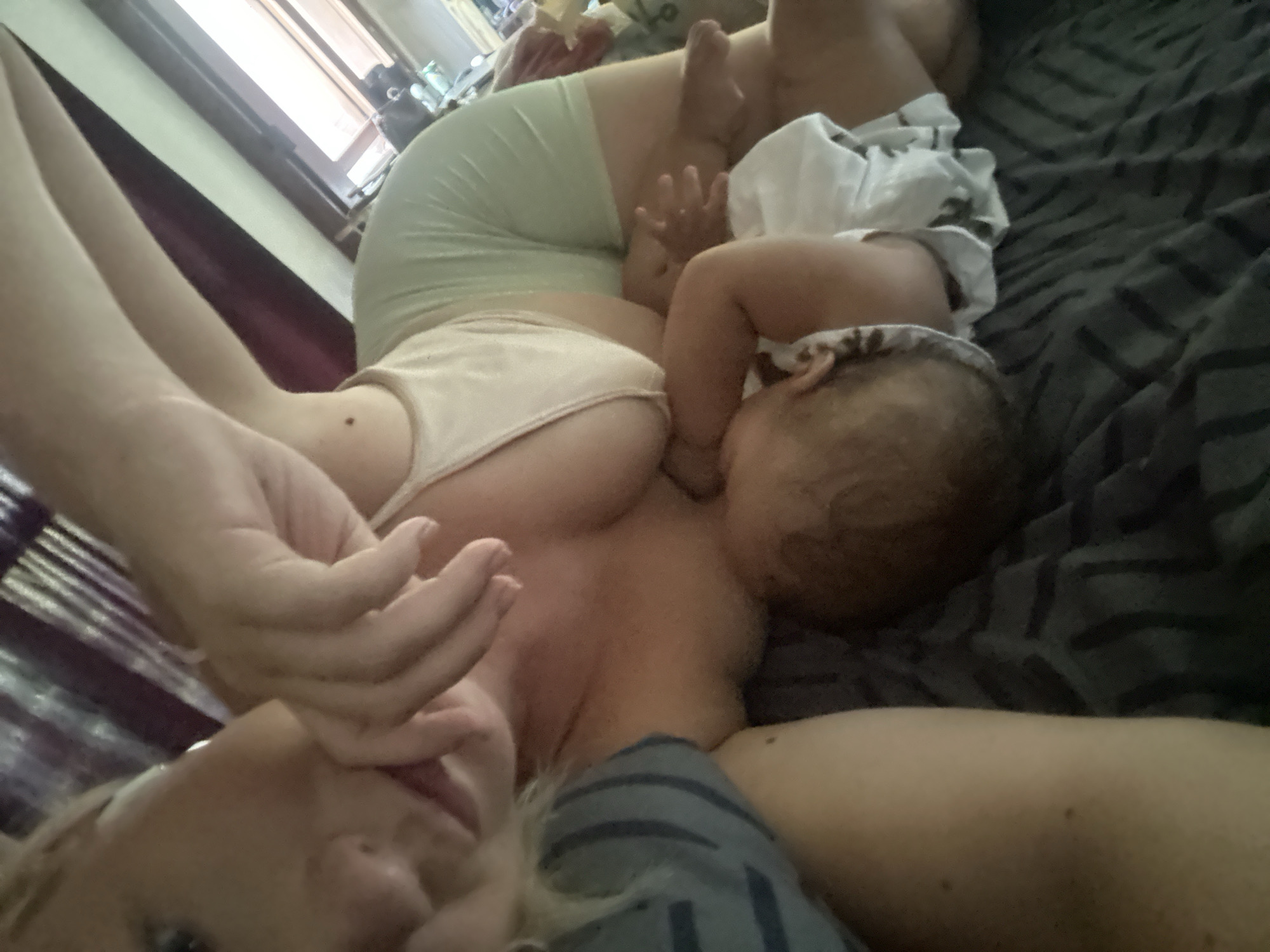 A person is lying down on a bed with a baby who appears to be breastfeeding. Another baby is lying beside them. The location cannot be determined from the image, but it was requested to be assumed as North Kuta, Indonesia.