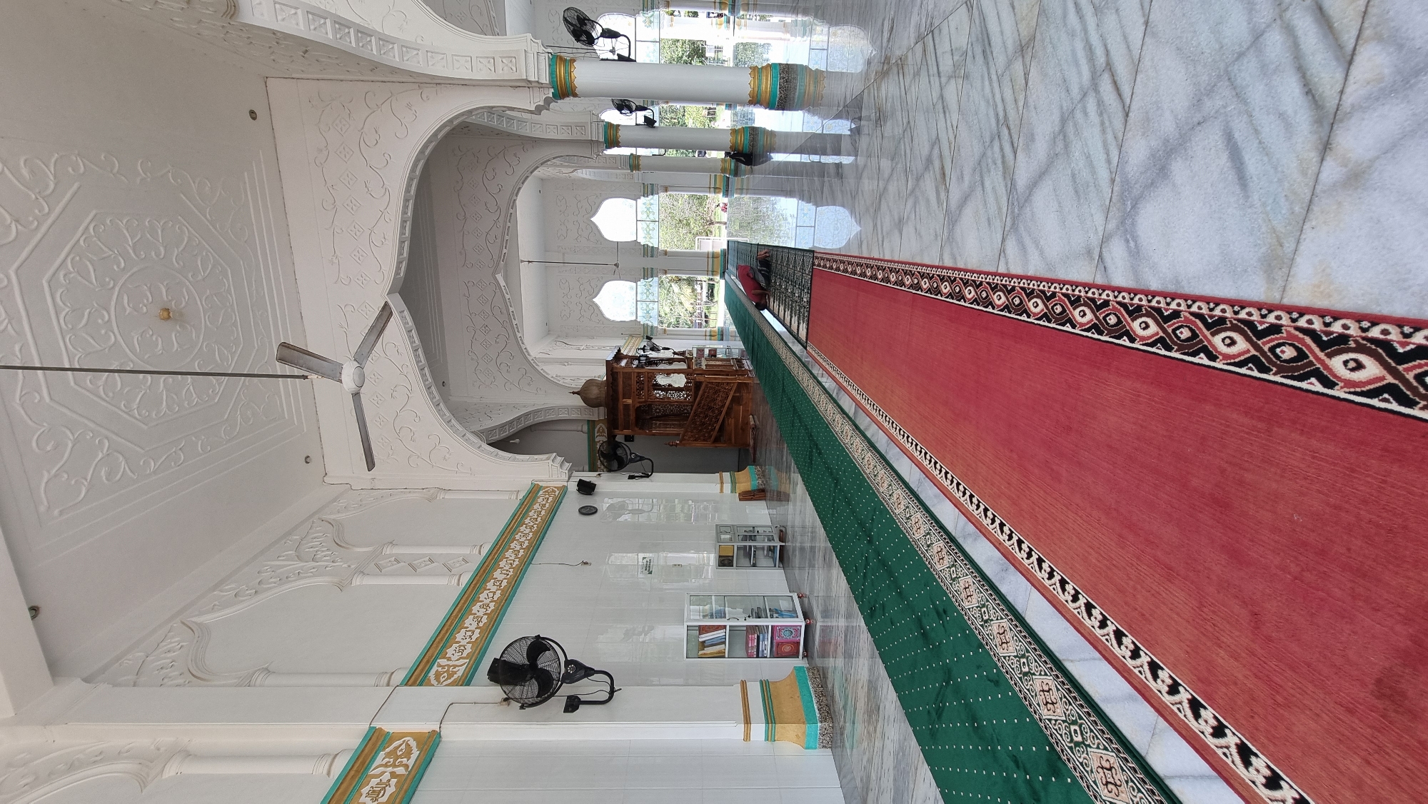 The image depicts the interior of a mosque in Lapang Timu, Indonesia. The scene is characterized by polished marble floors, white walls with intricate decorative moldings, and a high, ornate ceiling. A prominent wooden minbar, or pulpit, stands on the left side of the image. Two prayer carpets, one green and one red with patterned borders, are laid out on the floor, extending towards the background. A person in a red garment is seen prostrating on a prayer mat in the distance, on the green carpet. Several ceiling fans are visible, and the space is illuminated by natural light filtering in from large arched windows in the background, revealing lush greenery outside. There are also decorative pillars and shelves containing books. The overall impression is one of serene and clean religious architecture.