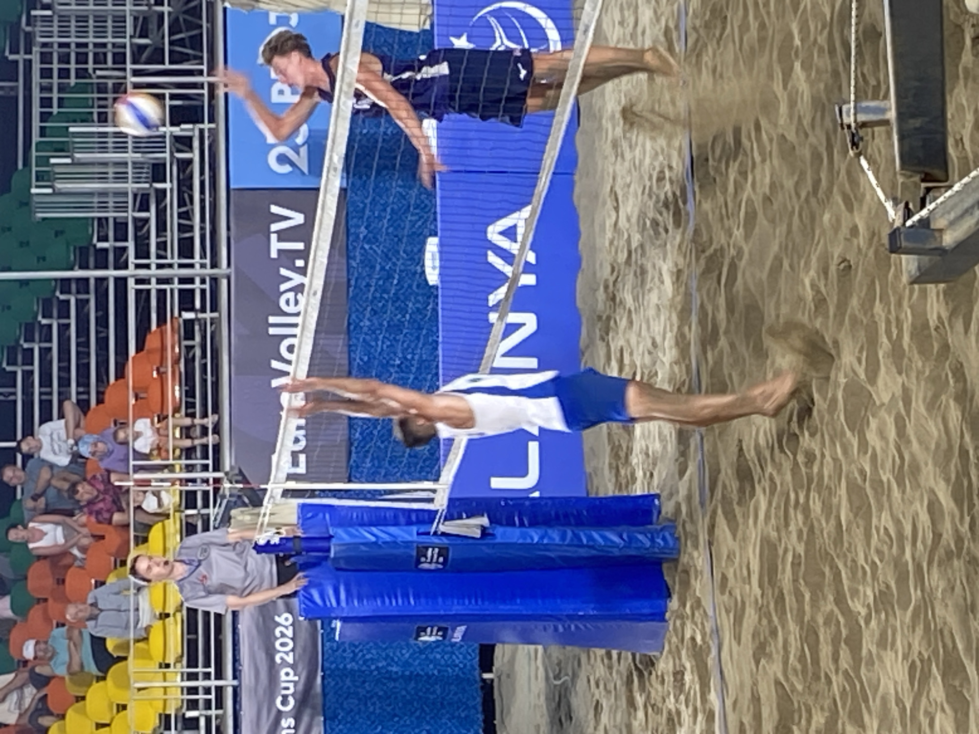 A beach volleyball match is captured at an outdoor court in Alanya, Turkey, likely during an evening event, indicated by artificial lighting. The scene features two male players, a female referee, and numerous spectators.
In the foreground, a male player wearing a white tank top with blue sides and blue shorts is airborne, having just jumped from the sand, with his back partially turned towards the viewer. He appears to be initiating an attacking maneuver. Directly opposite him across a white volleyball net, a second male player in a navy blue tank top and matching shorts is also airborne, with his arms raised, having just interacted with a multicolored volleyball that is suspended mid-air above and slightly behind him. Sand particles are visibly displaced by the players' movements on the court.
To the left of the net, a female referee stands on an elevated platform, wearing a grey polo shirt with a badge, dark shorts, and a microphone headset. She maintains a neutral expression, observing the play. Blue padded structures cover the net posts on her side of the court.
In the background, a series of blue banners display event branding, including "EuroVolley.TV", "ALANYA" (partially visible), and "ans Cup 2026". Behind these banners, tiered stadium seating with yellow, orange, and green seats is filled with numerous spectators of varying ages, casually observing the match. The overall environment suggests a professional sports event. A dark metal structure with ropes is visible in the bottom right corner of the frame.
