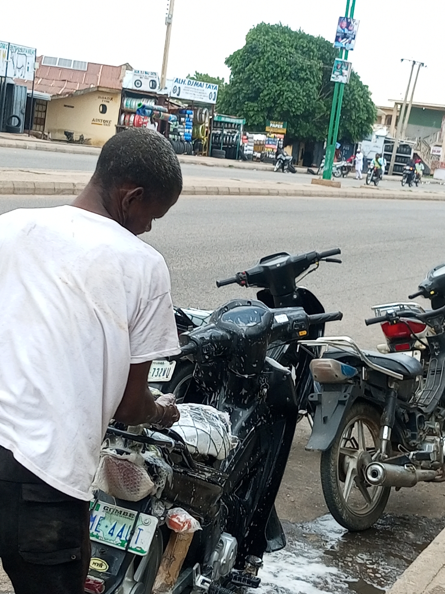 This is a forensic, matter-of-fact description of a scene in Arawa, Nigeria. A person, seen from behind and from the shoulder down, is wearing a dirty white t-shirt and dark pants. They are standing next to a black motorcycle, which is covered in soap suds, and appears to be in the process of being washed. The motorcycle has a license plate that reads "GOMBE ME 44LT". Another black motorcycle is partially visible to the right, and a third to the left. In the background, there are shops displaying tires and other goods, with signage that includes "ALH. DJMAI TAYA" and "ROADX TYRES". Several other motorcycles and people are visible in the distance, suggesting a busy commercial area. The ground is wet and soapy, indicating the washing activity. The overall environment is an urban street scene.