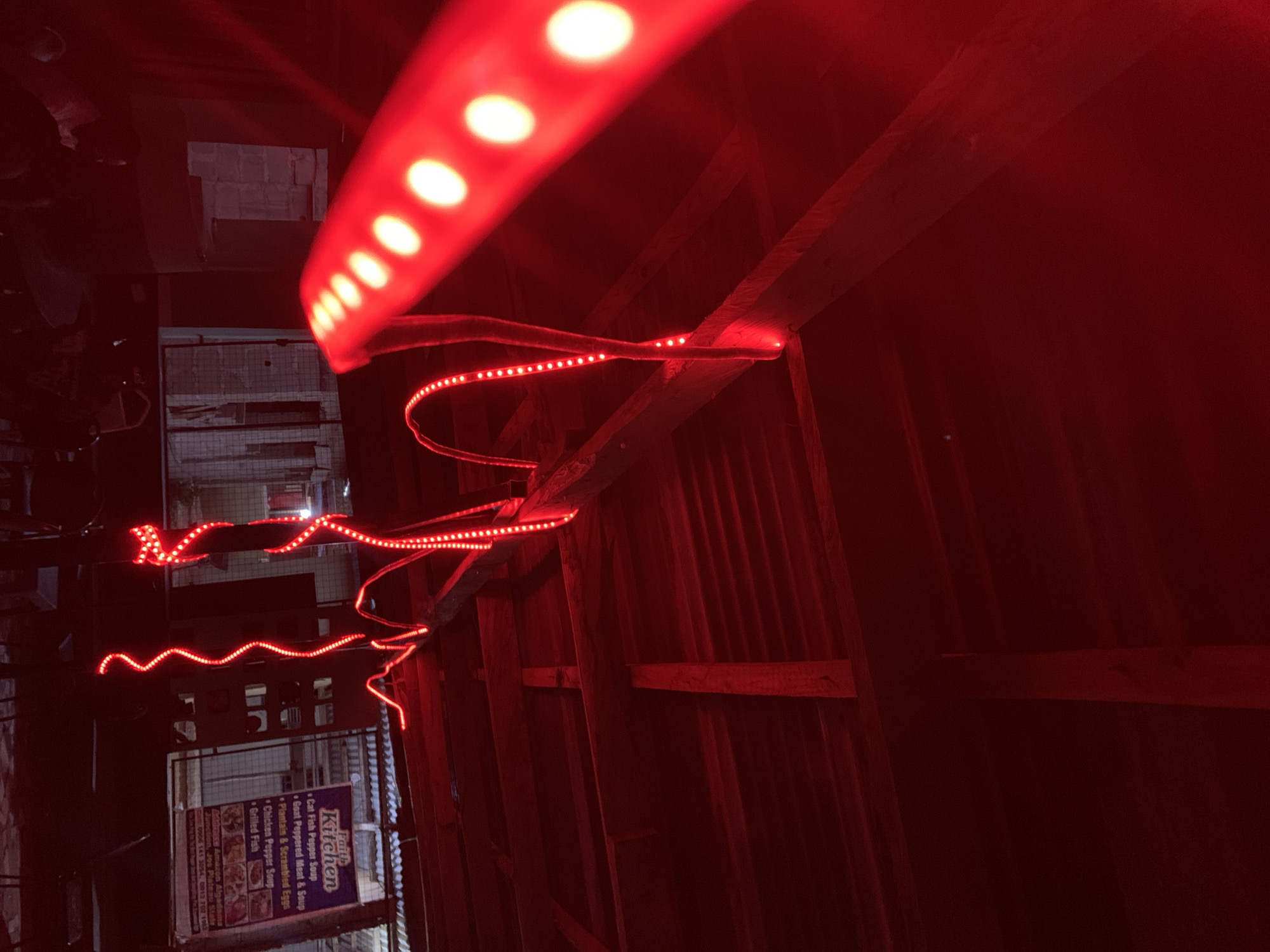 The image shows a building facade illuminated by red LED lights. The building seems to be constructed of corrugated metal sheets and wooden beams. In the lower left corner, a sign for "Faith Kitchen" is visible, listing menu items such as "Cat Fish Pepper Soup," "Goat Peppered Meat & Soup," and "Grilled Fish." The sign also mentions the location as "Amazon Algadama Junction, Jos Plateau State" in Nigeria, with contact numbers 0906 141 3054 and 0813 702 1962. The environment appears to be a dimly lit street at night.