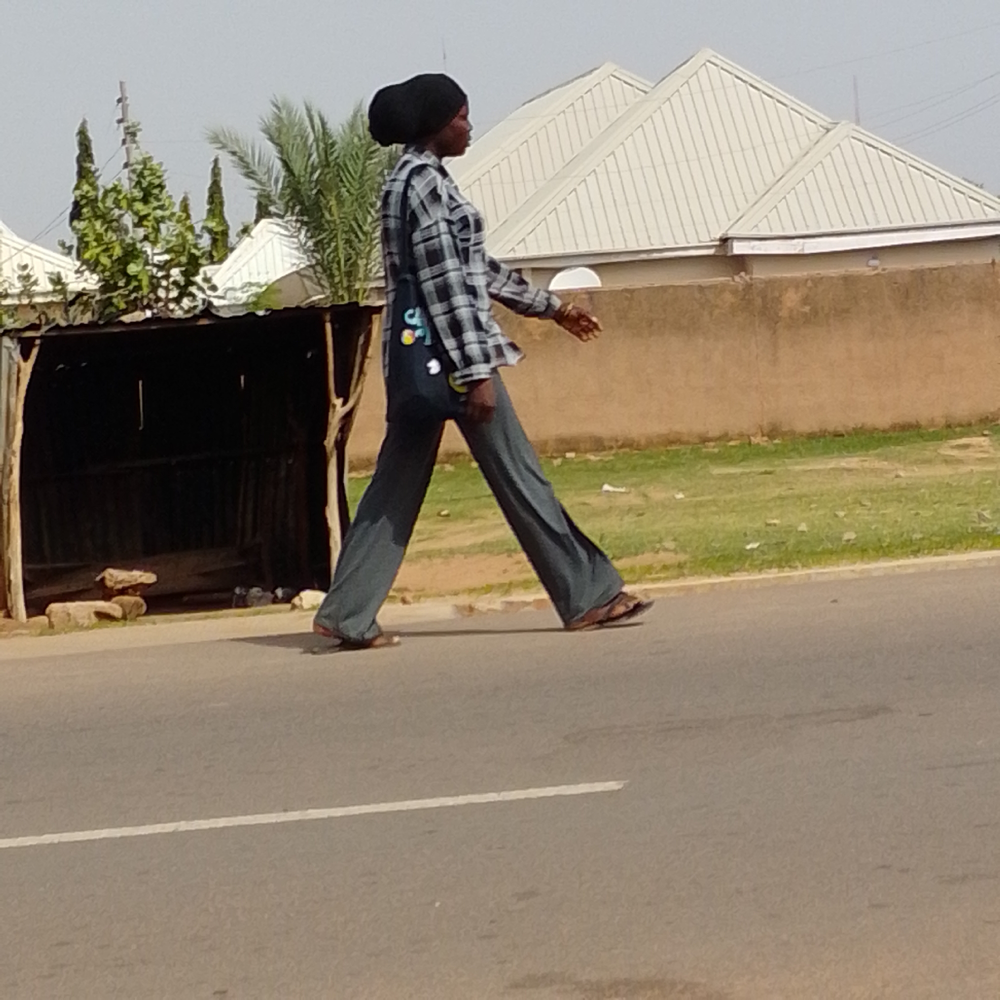A full-bodied person is walking on a paved road in Gombe, Nigeria. The person is wearing a black hijab, a grey and white checkered long-sleeved shirt, and dark grey flared pants. They are also wearing brown sandals. The person is walking from left to right across the frame. In the background, there are buildings with white roofs and brown walls, trees, and a dark, open structure that appears to be a shelter or stall. The sky is a pale blue. The road has a white line painted on it, indicating a lane divider.