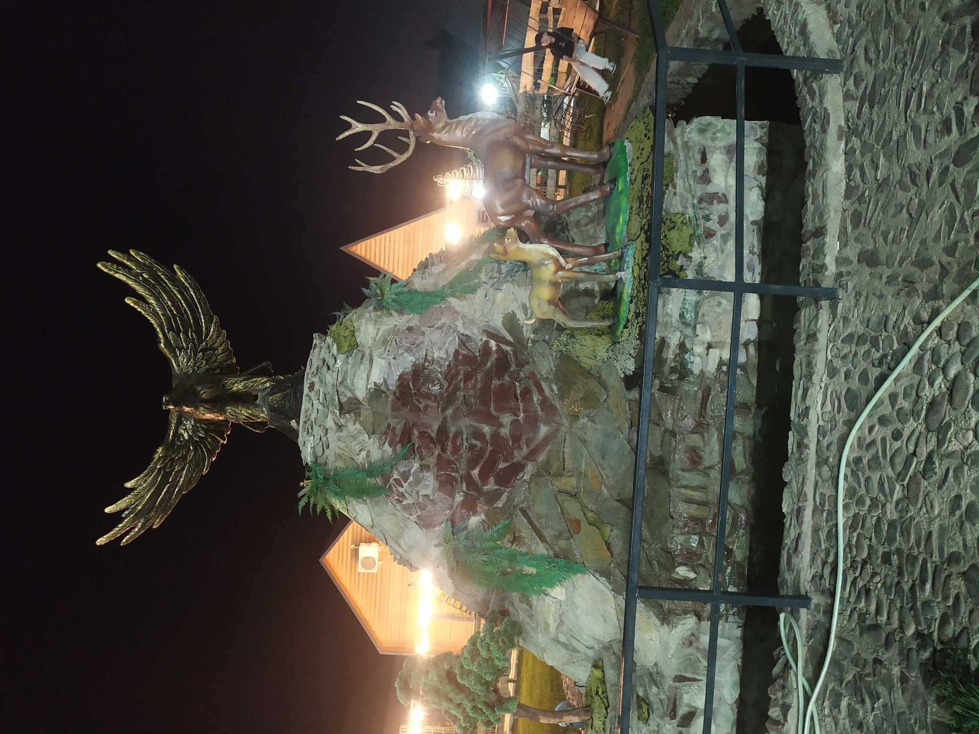 The image depicts a nighttime scene in Sosnovka, Kyrgyzstan, featuring an elaborate outdoor decorative fountain and landscaping. Dominating the central area is a large, multi-tiered rock structure, likely artificial, adorned with green, fern-like decorations. Atop this structure is a striking statue of a golden eagle with outstretched wings. At the base of the rock formation, two deer statues are positioned: a larger stag with an impressive rack of antlers and a smaller fawn.
The surrounding environment includes a building with a distinctive triangular roof, illuminated from within and by external lights, suggesting it could be a restaurant, cafe, or event space. To the left, a miniature, conical evergreen tree is visible. A dark, water-filled channel or moat runs in front of the rock structure, bordered by a black metal railing. The ground is paved with irregular stones, and a white hose lies on the paving stones.
To the right of the fountain, a wooden swing set is partially visible. A person, dressed in light-colored pants and a dark shirt, is seated on one of the swings, looking towards the left. The overall ambiance is enhanced by artificial lighting, creating a warm and inviting atmosphere despite the darkness of the night. The scene suggests a public recreational area or a hospitality venue.